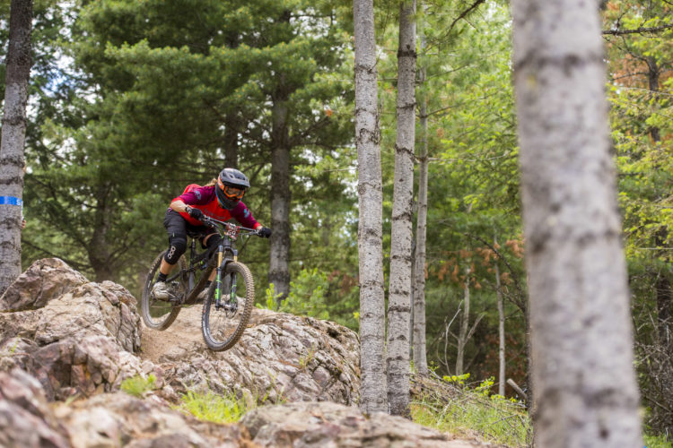 A mountain biker dressed in a red and black jersey navigates a rocky trail surrounded by tall trees in a dense forest. The rider is airborne over a large rock, showcasing skill and agility on their bicycle against a backdrop of greenery.