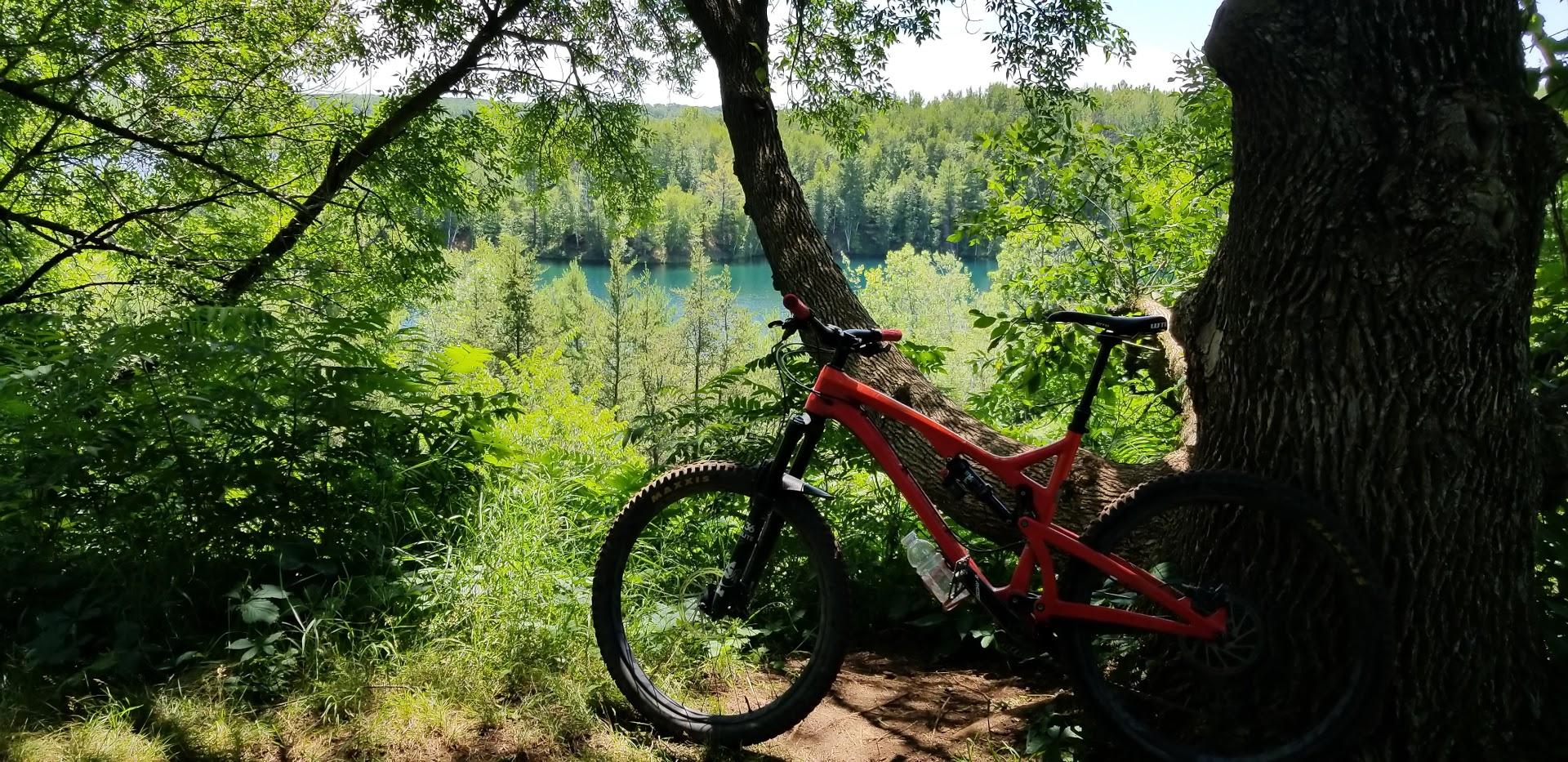 A red mountain bike is leaning against a tree, surrounded by lush green foliage. In the background, a serene body of water is visible, framed by trees and vegetation, depicting a peaceful outdoor scene ideal for biking or nature exploration. Cuyuna Lakes mountain bike trail.