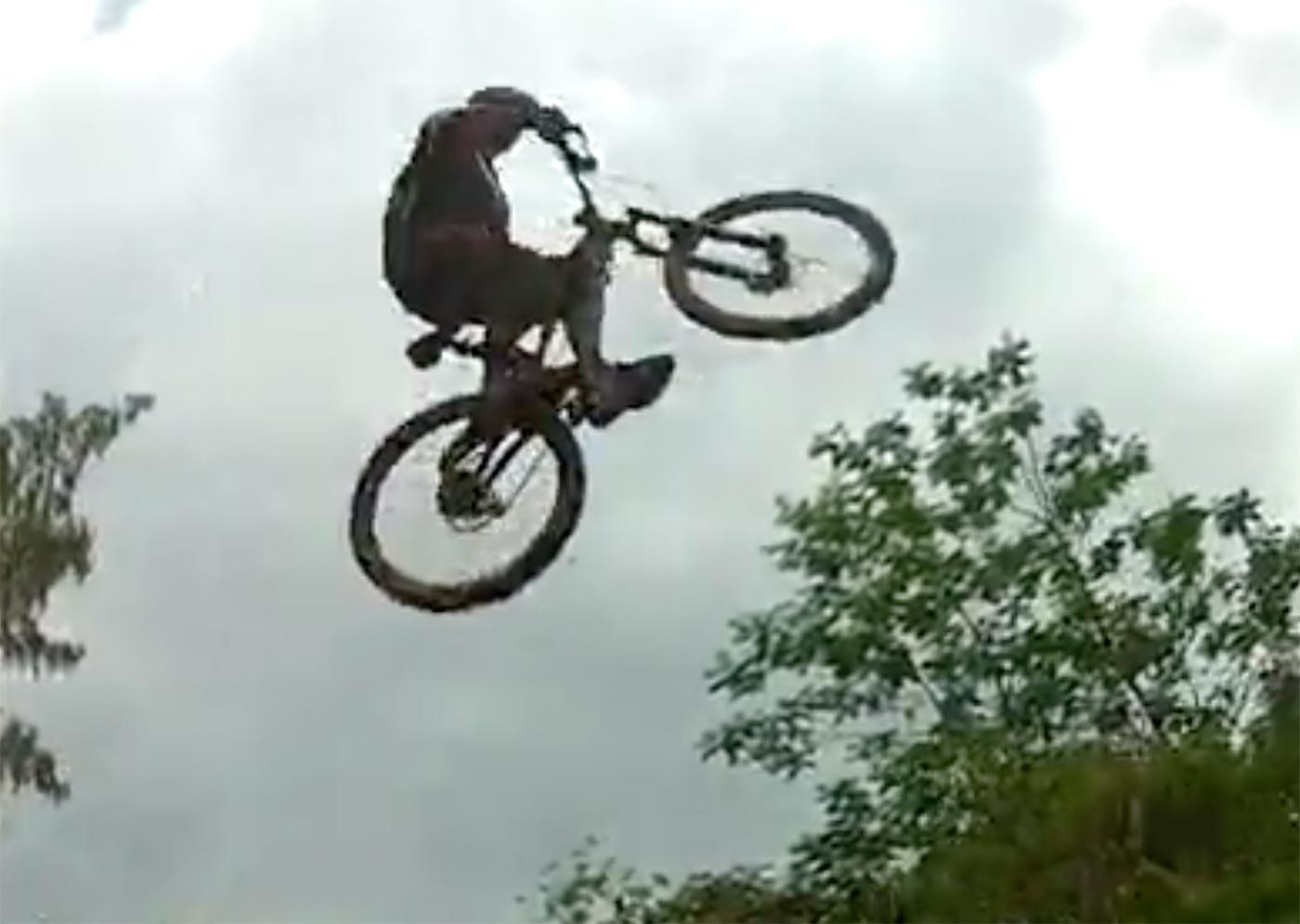 A mountain biker performs a jump in mid-air, showcasing an impressive aerial maneuver against a cloudy sky, with trees visible in the background.