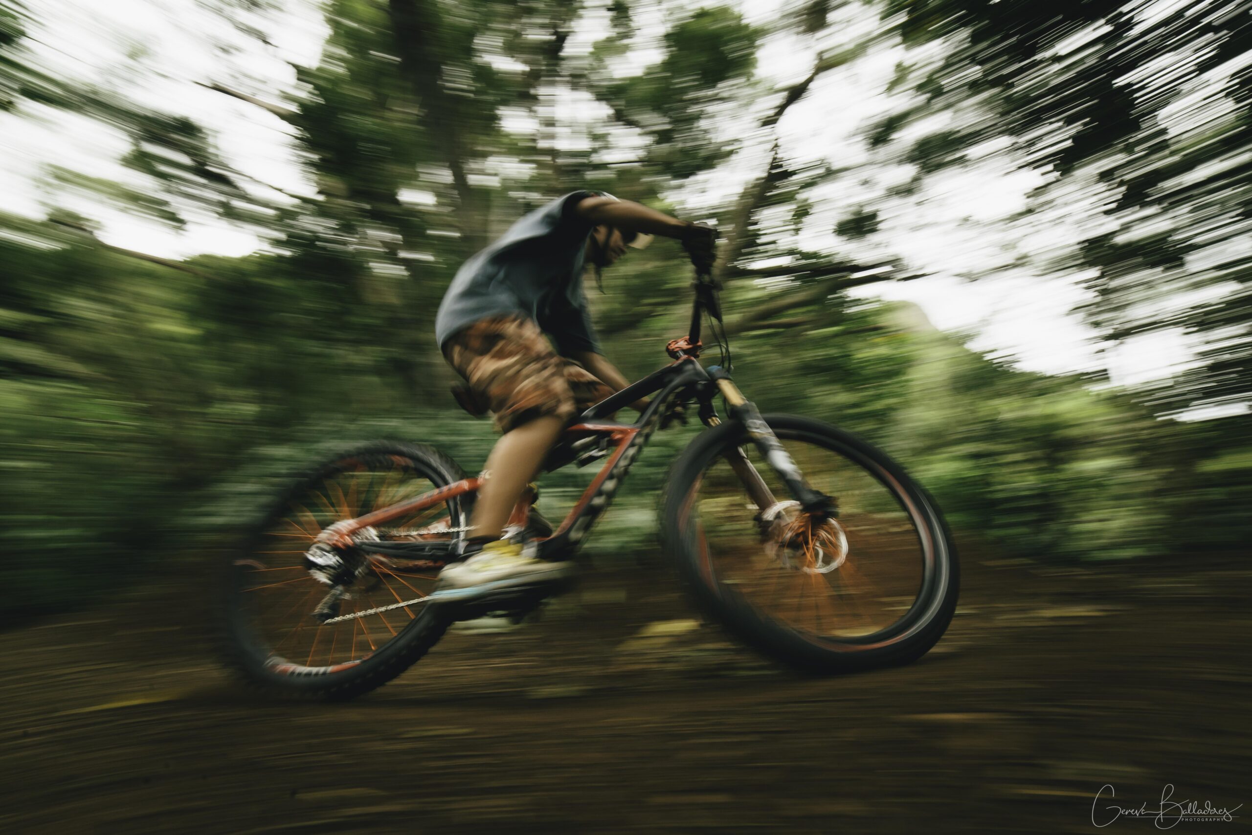 A cyclist riding a mountain bike at high speed through a wooded area, captured with motion blur to emphasize movement and excitement. The background shows a lush green environment, creating a dynamic sense of adventure.