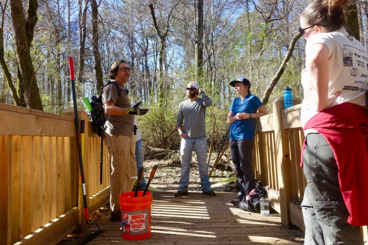 A group of four people engaged in conversation on a wooden bridge in a wooded area. They are surrounded by trees and greenery. Tools are resting against the bridge and in a bright orange bucket labeled for outdoor use. The individuals vary in attire and appear to be discussing a project or activity related to the outdoors.
