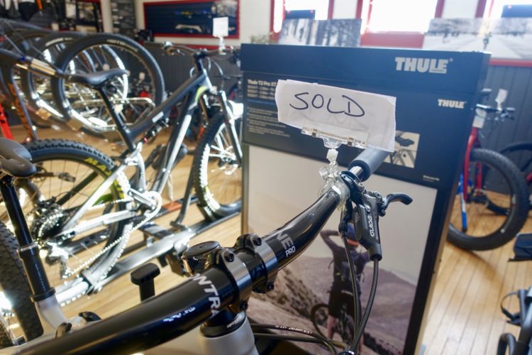 A close-up view of a bicycle handlebar with a "SOLD" sign attached, showcasing the bike's model details. In the background, other bicycles and a display board with product information can be seen, indicating a bike shop setting.