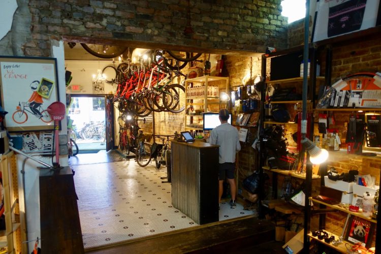 Interior view of a bicycle shop featuring a man at a wooden counter using a computer. The walls are adorned with bicycles hanging from the ceiling and shelves stocked with cycling accessories and gear. A decorative poster promoting cycling decorates the entrance area, while the floor has a patterned tile design. The shop has a warm, inviting ambiance with good lighting.