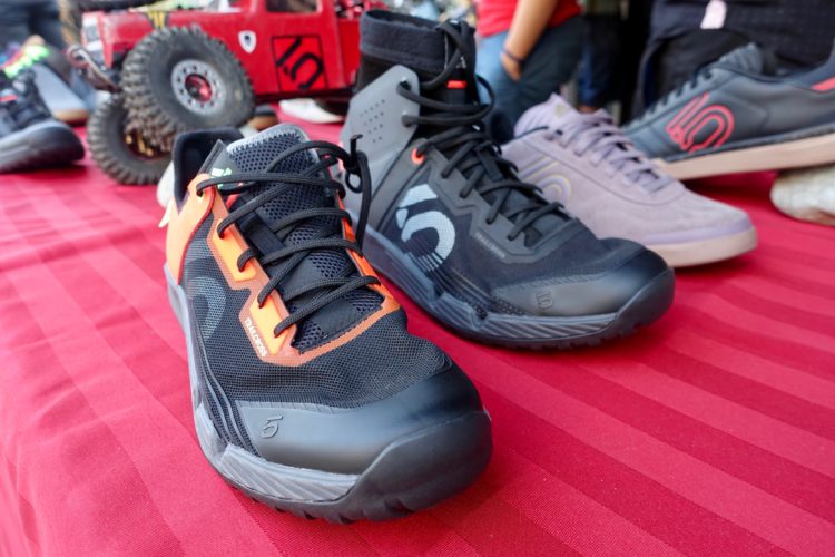 A close-up view of two pairs of athletic shoes displayed on a red striped tablecloth. The left shoe features a black mesh design with orange accents, while the right shoe has a predominantly black design with a grey accent. In the background, a small toy vehicle is partially visible, along with other shoes.