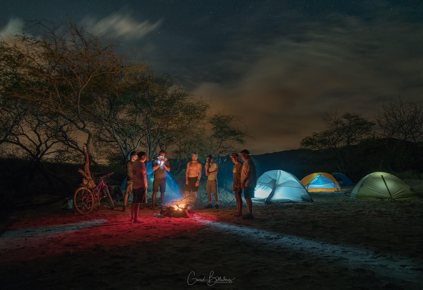 A group of people gathered around a campfire at night, with several illuminated tents in the background. The scene includes a bicycle resting nearby and a starry sky above, creating a warm, adventurous atmosphere.