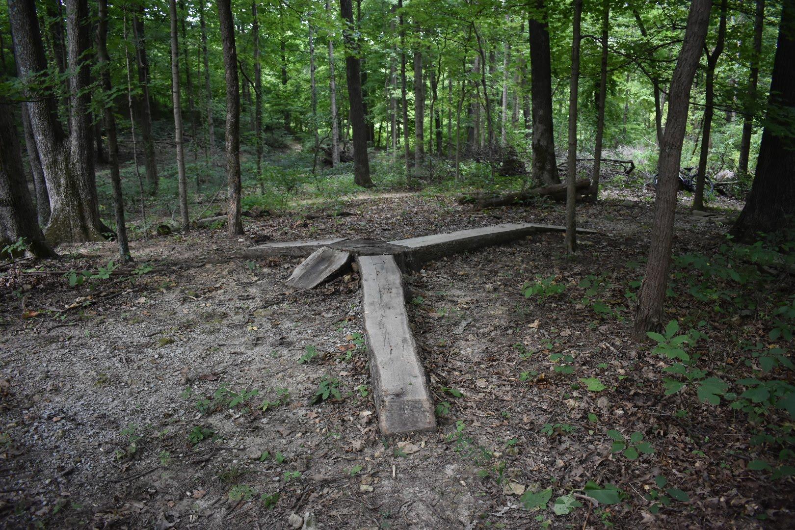 A wooden structure shaped like an arrow pointing in multiple directions, situated in a forested area with green foliage and trees surrounding it. The ground is covered in dirt and scattered leaves. Saunder Springs mountain bike trail.