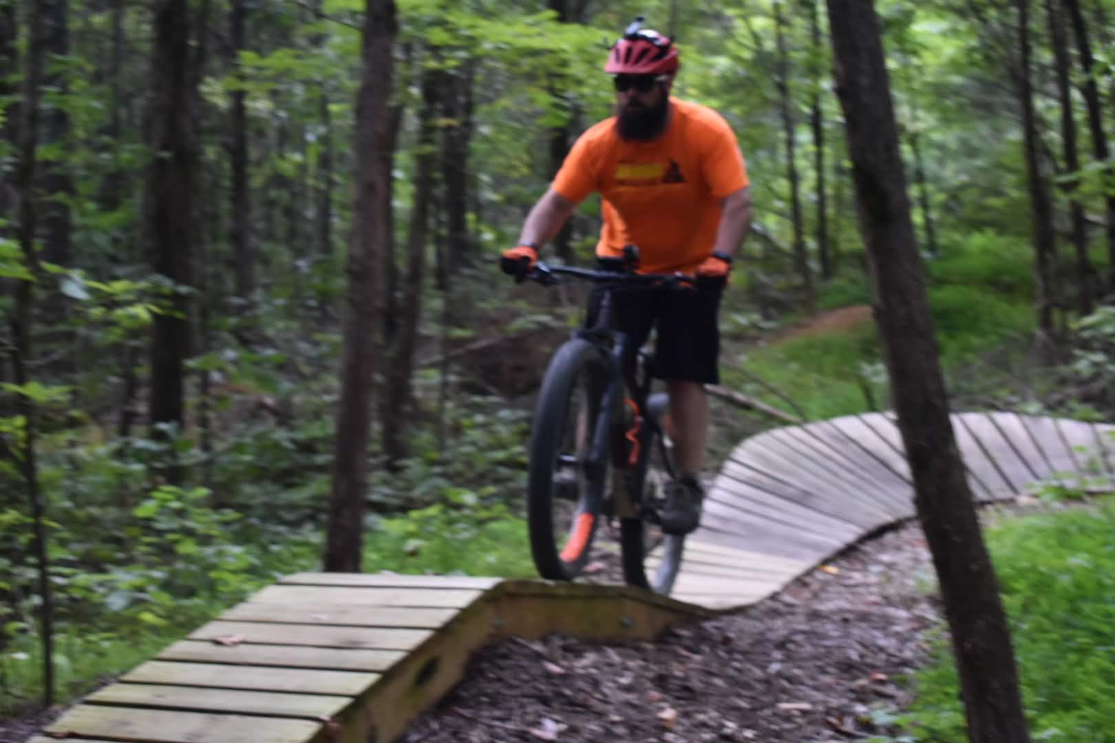 A person riding a mountain bike on a curved wooden trail in a lush, green forest. The rider is wearing an orange shirt, black shorts, and a helmet. The scene captures the dynamic action of biking along the trail surrounded by trees. Saunder Springs mountain bike trail.