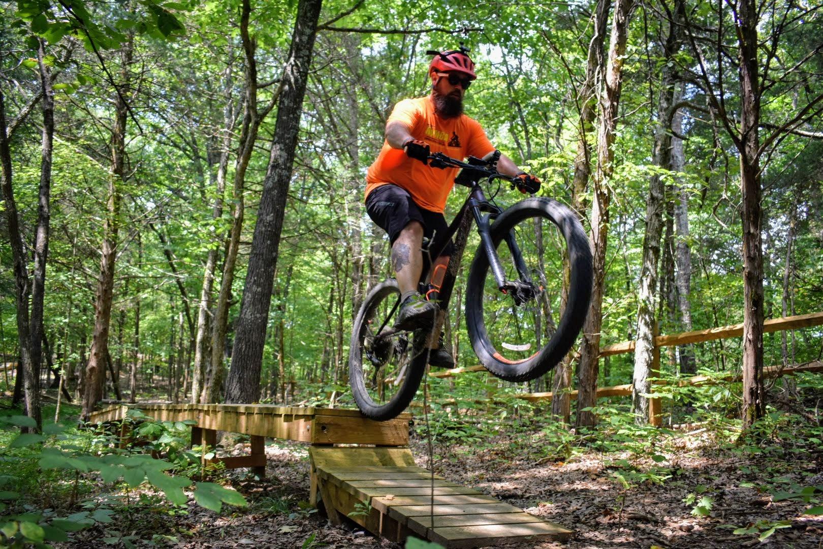 A mountain biker in an orange shirt and helmet is airborne over a wooden bridge in a lush green forest, surrounded by trees and foliage. The bike has wide tires, and the rider is focused on maintaining balance mid-jump. Sunlight filters through the leaves, creating a vibrant outdoor scene. Saunder Springs mountain bike trail.