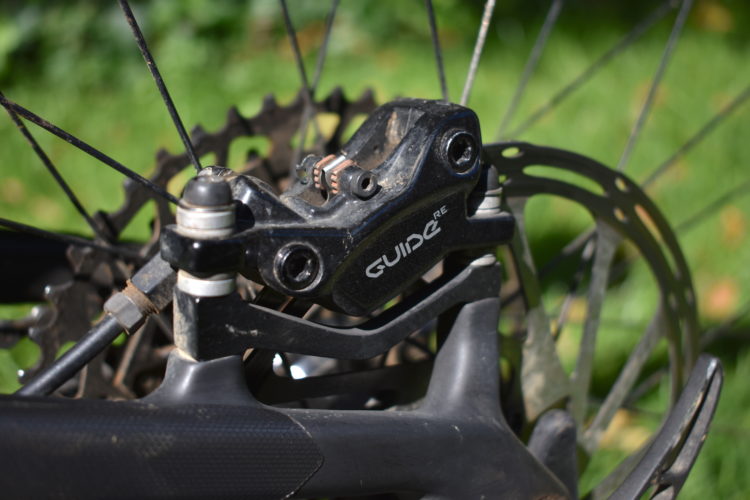 Close-up of a bicycle brake system, showcasing a black hydraulic brake caliper labeled "GUIDE RE" with detail on the disc rotor, brake lines, and components against a green grass background.