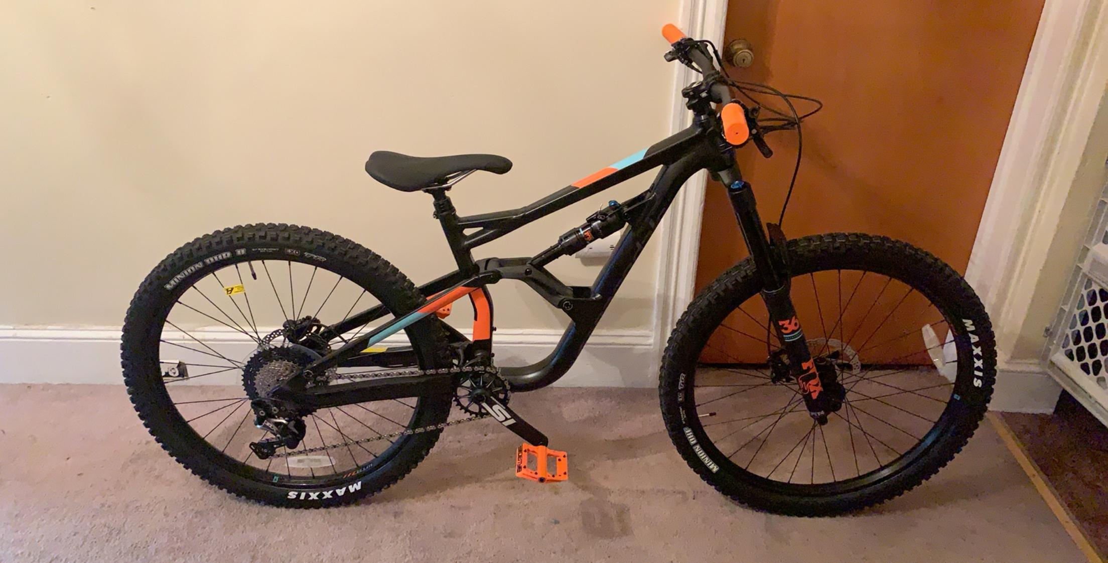 Cannondale Jekyll: Mountain bike with a black frame featuring colored accents, prominent tires with tread suited for off-road riding, and a sturdy build. The bike is positioned indoors against a wall with a brown door in the background.
