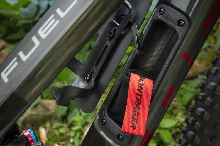 Close-up of a bicycle frame featuring a water bottle cage and storage compartment. The frame is dark gray with the word "FUEL" printed on it, and the storage compartment contains a black item with an orange strap labeled "BONTRAGER." The background includes green foliage and dirt, indicating an outdoor environment.
