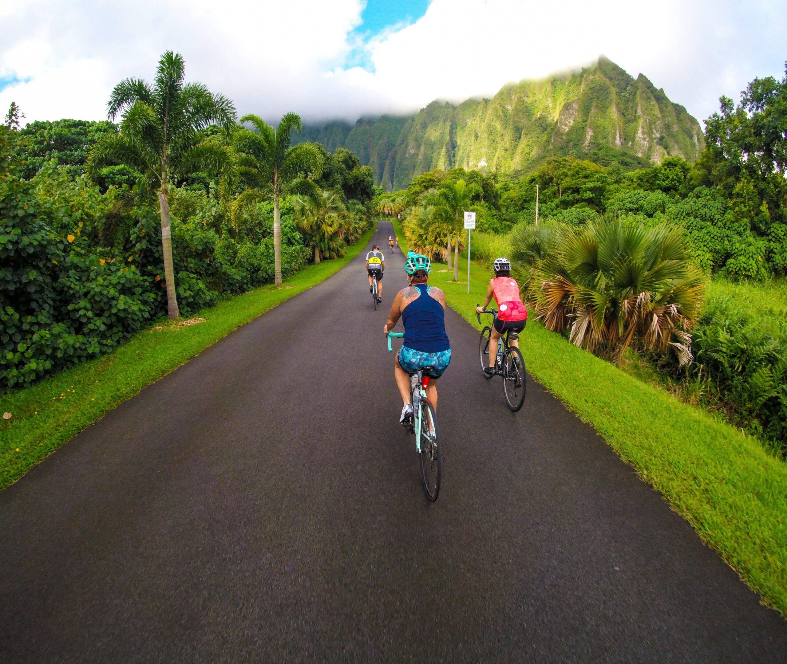 A group of cyclists riding along a paved road surrounded by lush greenery and towering mountains in the background. The sky is partly cloudy, with hints of blue peeking through.
