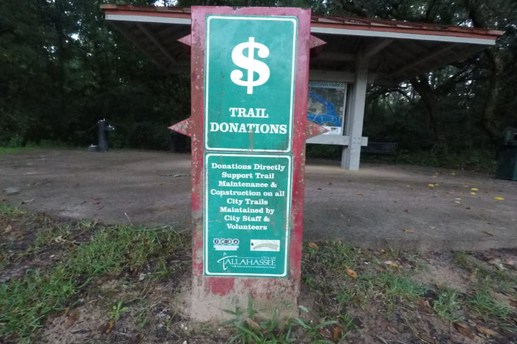Signpost indicating trail donations, featuring a green background with white text. The sign states "Trail Donations" and explains that donations support maintenance and construction of city trails, managed by city staff and volunteers. A structure with a roof is visible in the background.