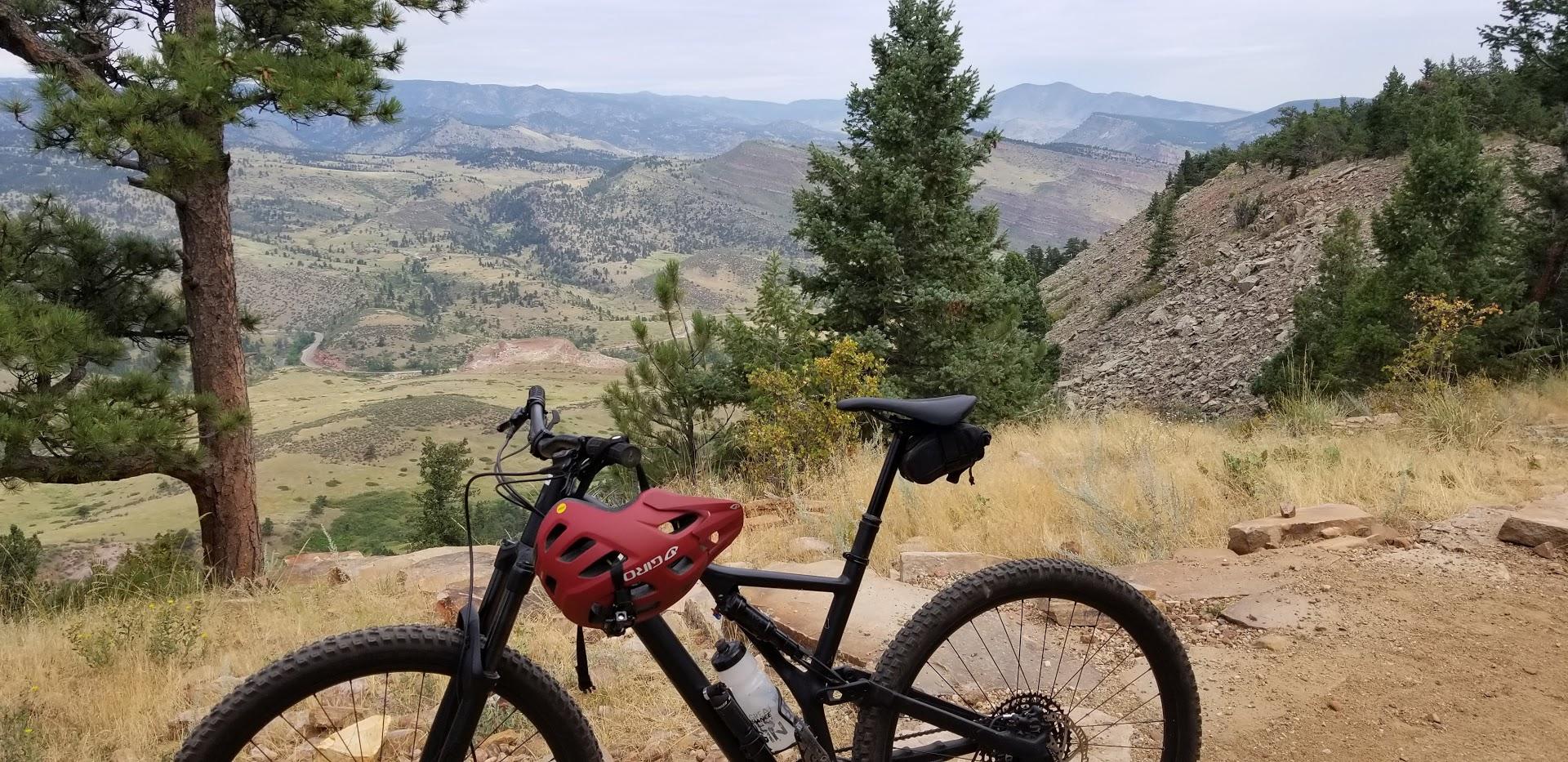 A mountain bike parked on a rocky trail, with expansive views of rolling hills, valleys, and distant mountains under a cloudy sky. Pine trees frame the scene, emphasizing the natural landscape. Heil Valley Ranch mountain bike trail.