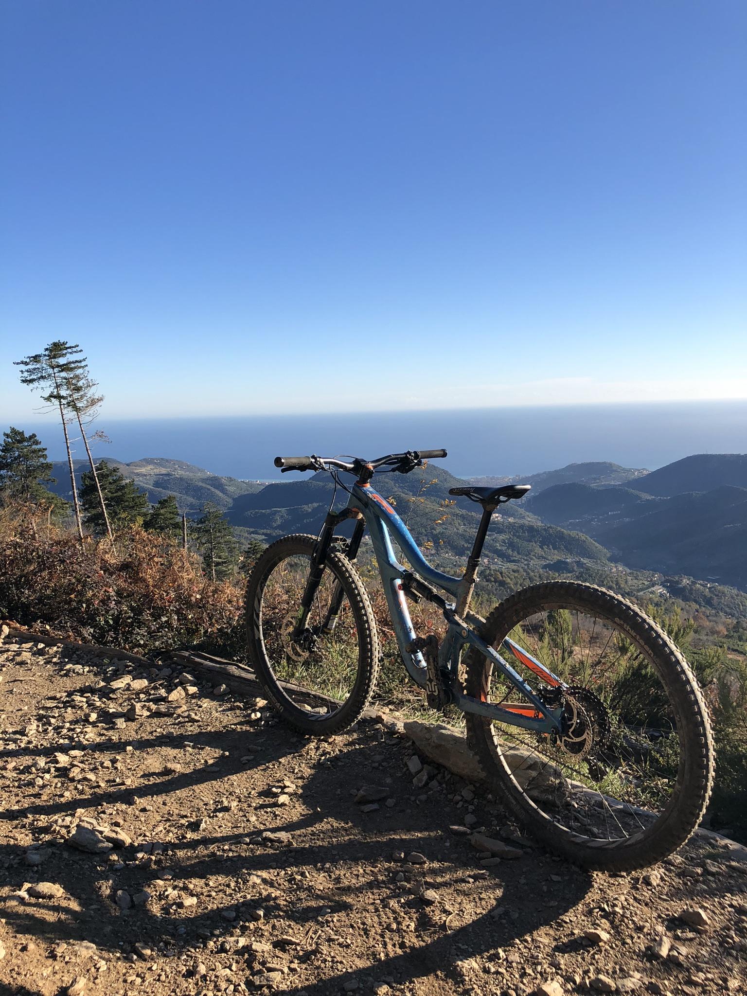 Ibis RipMo: Mountain bike resting on a rocky trail with a scenic view of hills and the ocean under a clear blue sky. Sunlight casts long shadows on the ground.
