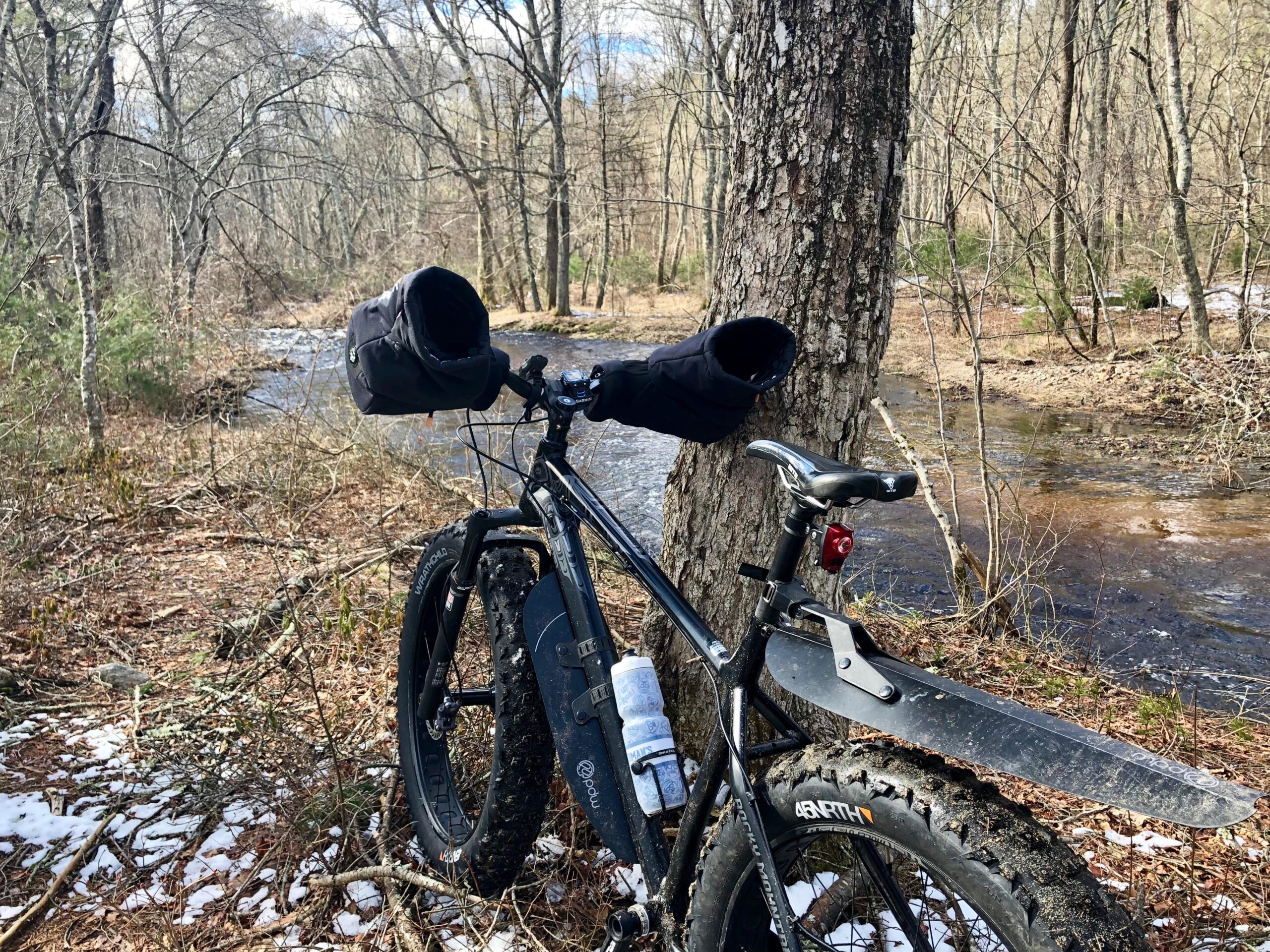 A mountain bike with winter gear, parked beside a flowing creek surrounded by bare trees and scattered fallen leaves. The bike features wide tires and a mudguard, with handlebar mitts for added warmth. Snow is visible on the ground in some areas.