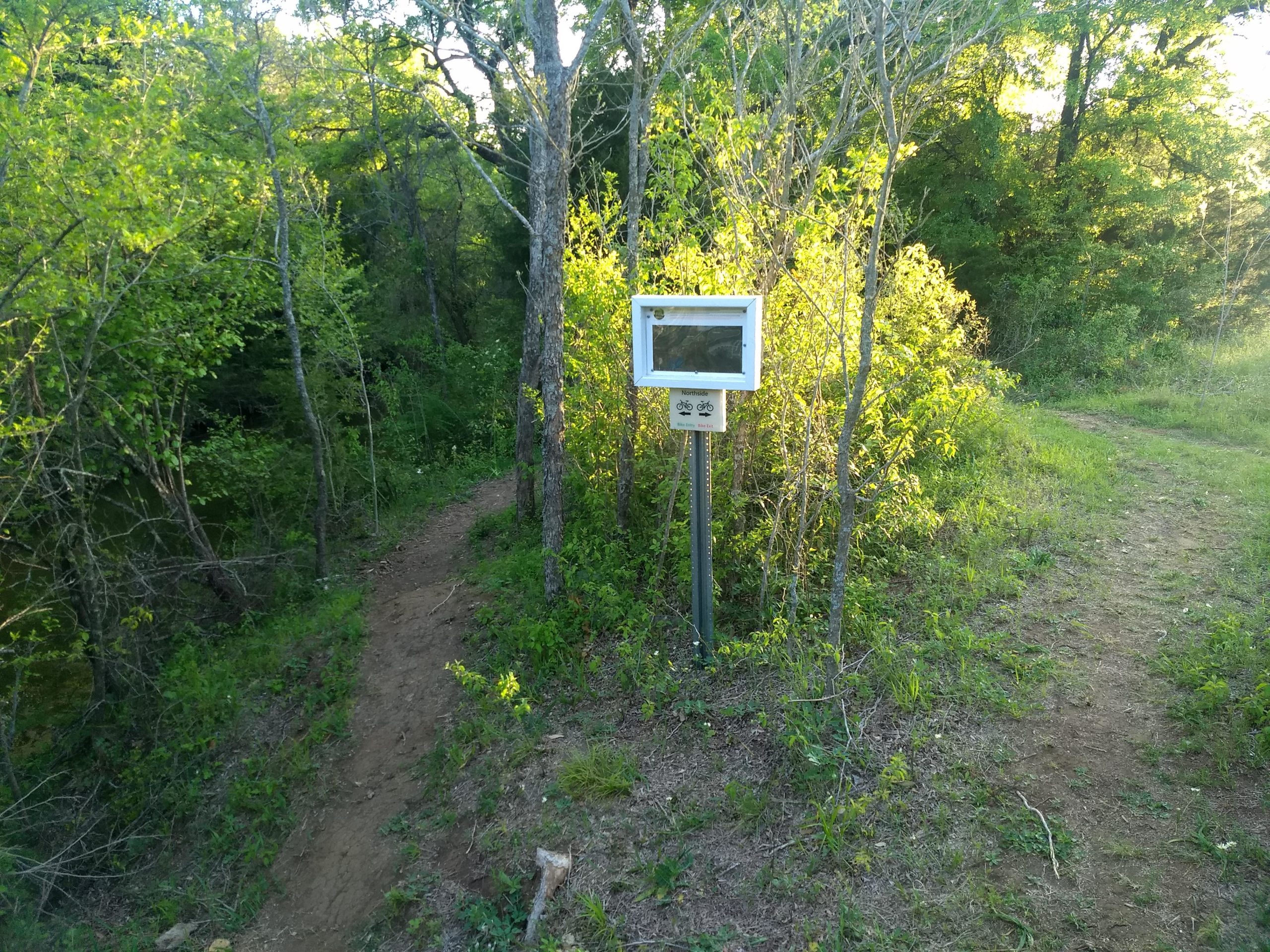 A narrow dirt path surrounded by greenery, leading through a wooded area. To the side, there is a white sign mounted on a post, which includes a graphic of a bicycle, indicating that the path is suitable for biking. The scene is bathed in warm sunlight filtering through the trees. Chisenhall trails mountain bike trail.