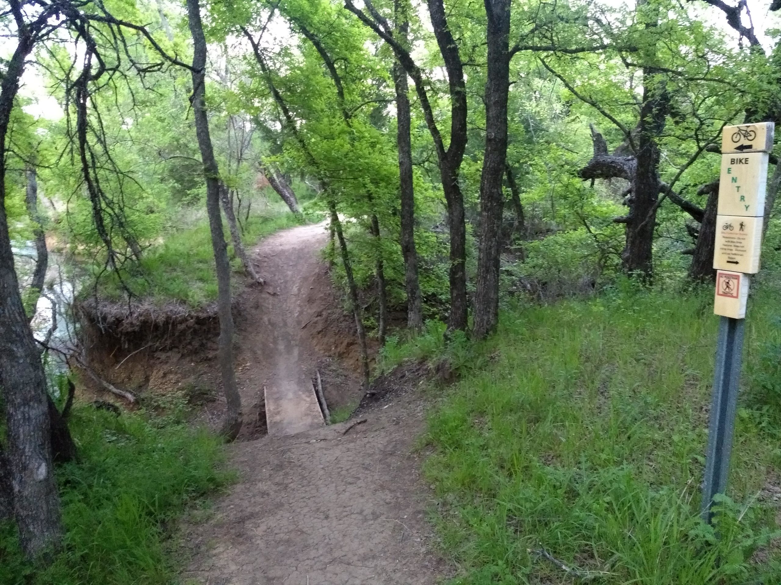 A narrow dirt trail winding through a lush green forest with trees on either side. A wooden bridge crosses a small dip in the terrain, leading to a path that curves to the left. To the right, a signpost indicates "Bike Entry" with additional information and guidelines for trail users. Chisenhall trails mountain bike trail.