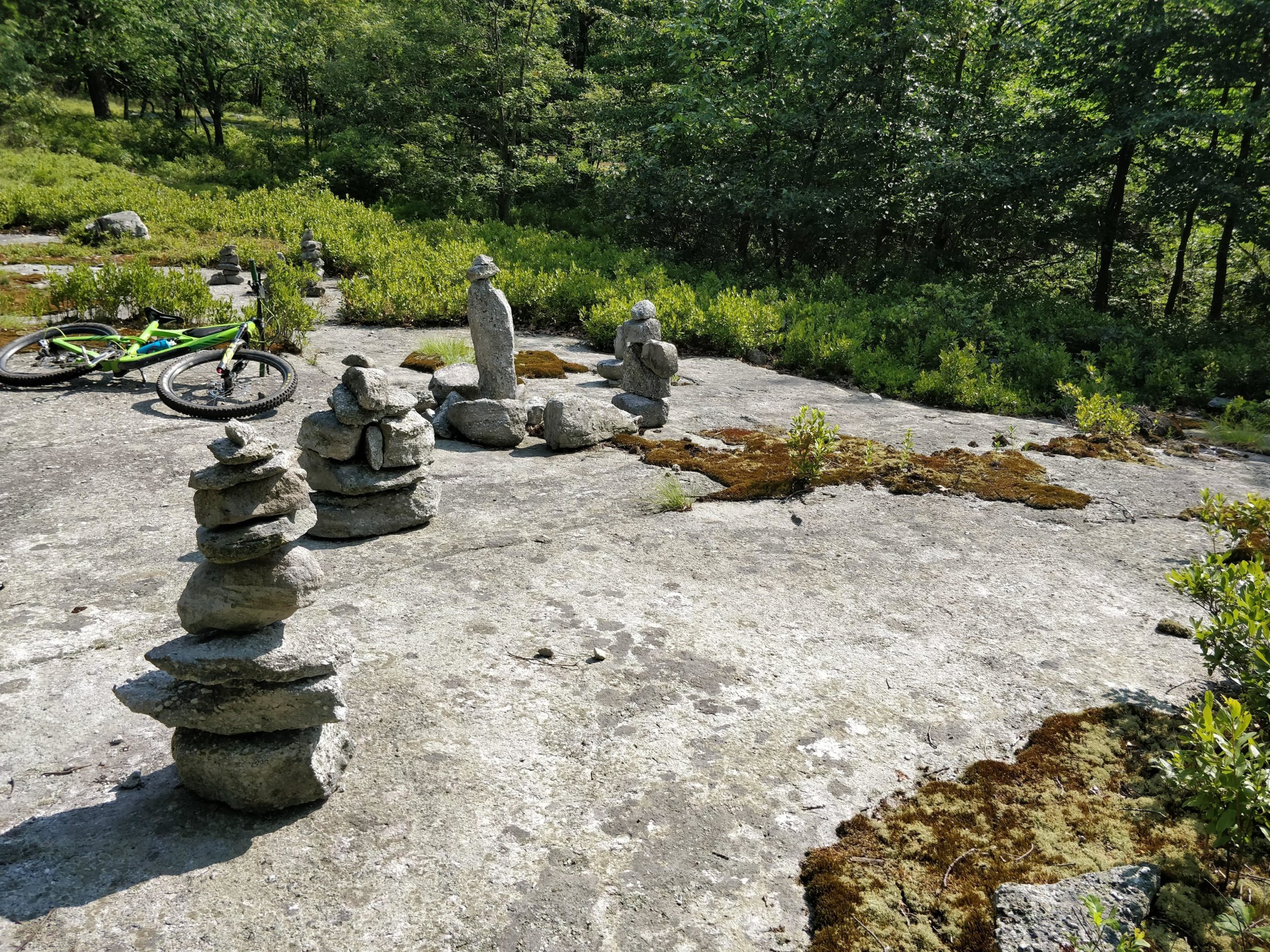 A serene outdoor scene featuring multiple stacked stone cairns on a rocky surface, surrounded by patches of greenery and moss. A green mountain bike is positioned nearby, partially in view, under a bright blue sky with trees in the background. Blueberry mountain bike trail.