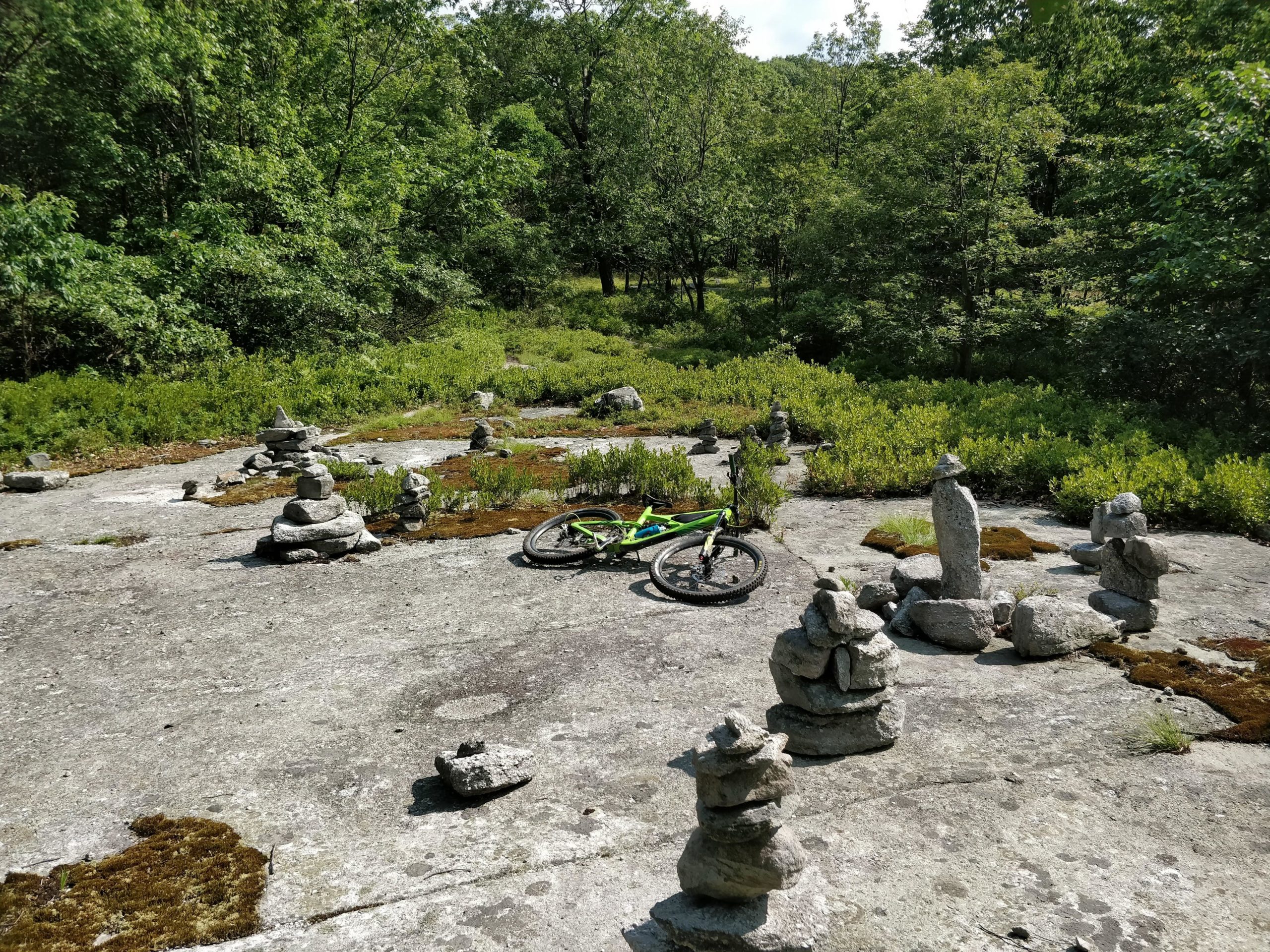 A mountain bike rests on a rocky surface surrounded by numerous stacked stone cairns, with lush green foliage and trees in the background under a clear sky. Blueberry mountain bike trail.