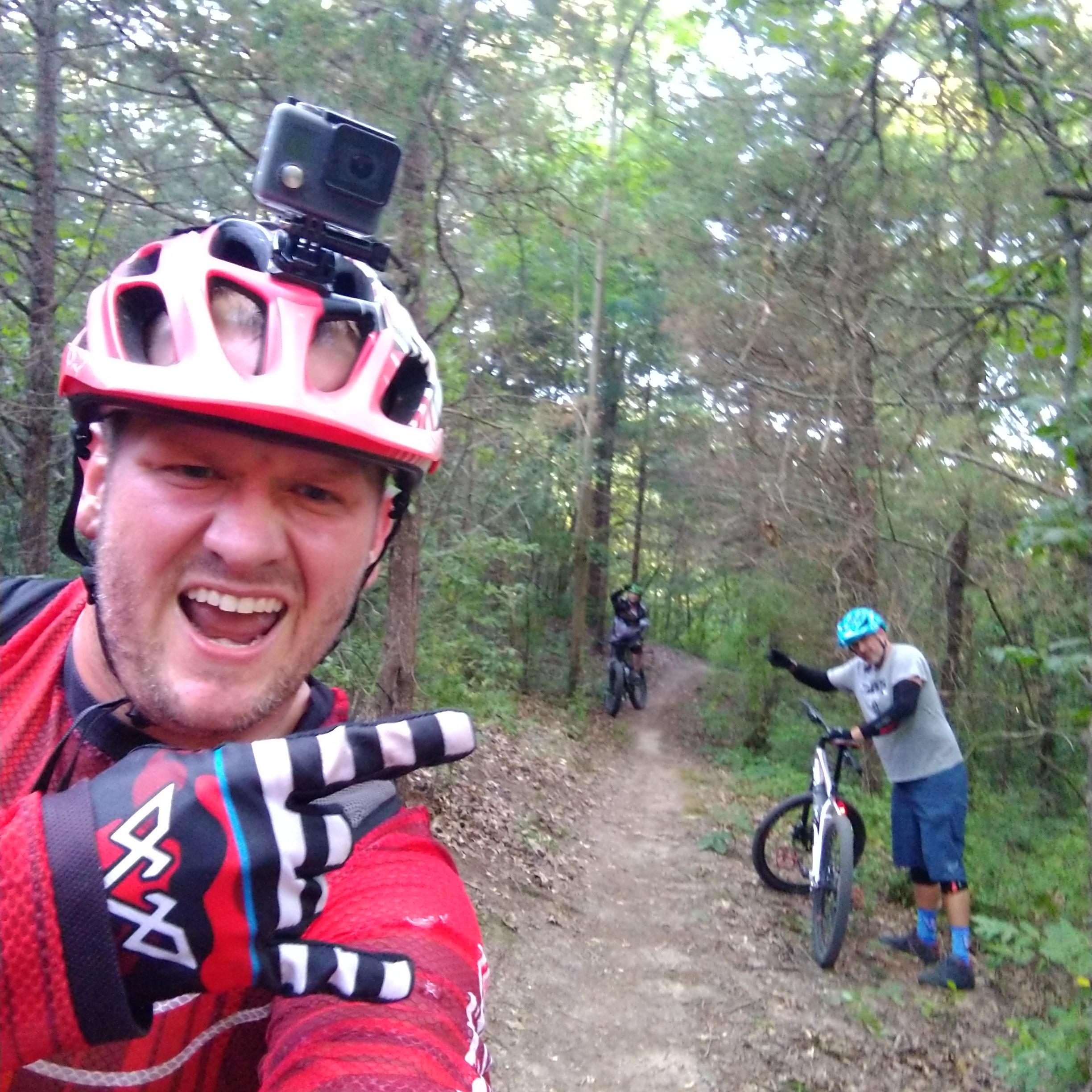 A group of three mountain bikers on a forest trail. The foreground features a smiling cyclist wearing a red helmet and gloves, making a hand gesture with excitement. In the background, two other riders can be seen, one waving while the other is positioned on the trail. The scene is surrounded by lush greenery, highlighting a sunny day outdoors. Brighton Rec Area mountain bike trail.
