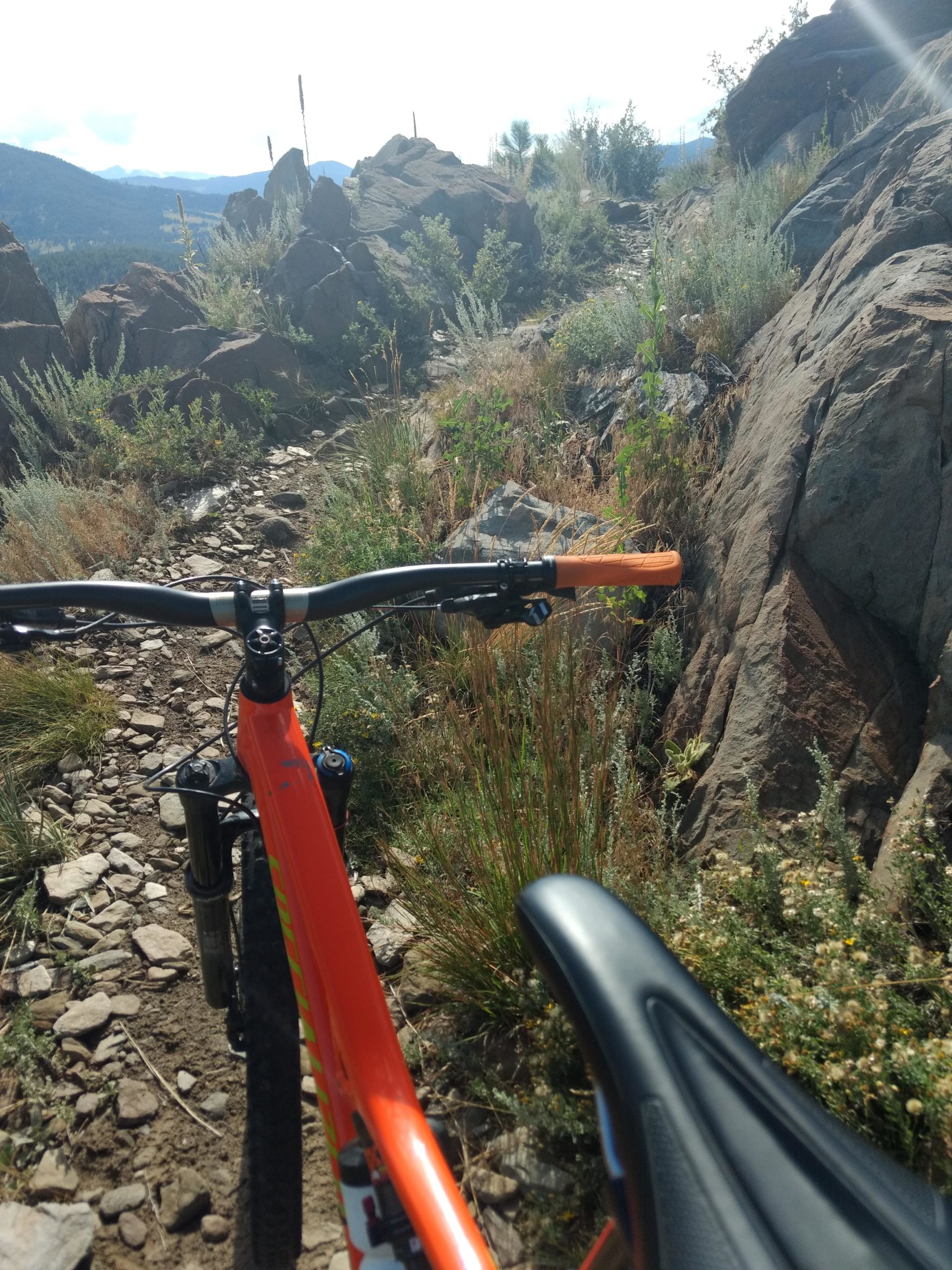 A close-up view of a mountain bike's handlebars and seat, overlooking a rocky trail surrounded by greenery and hills in the background, under a bright blue sky. Ginny Trail mountain bike trail.