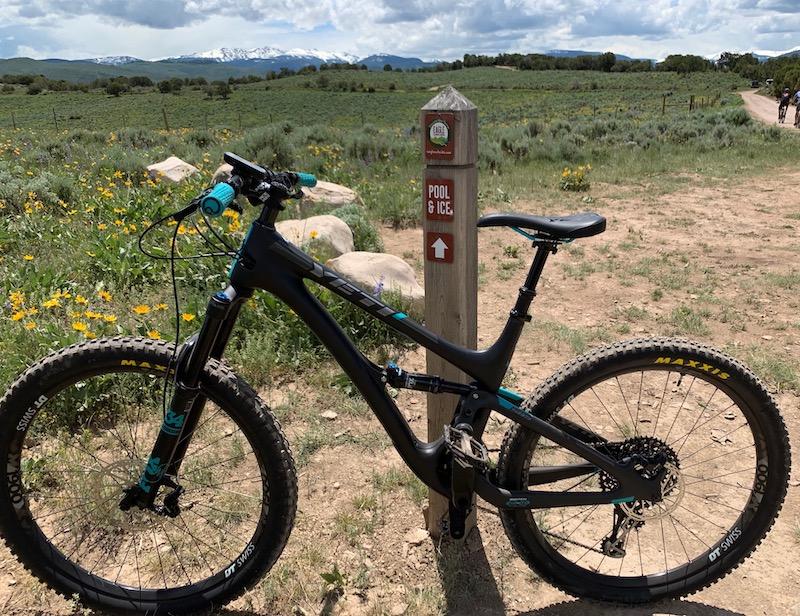Yeti SB5: A black mountain bike with turquoise accents leaning against a signpost in a grassy area, with wildflowers and mountains in the background under a cloudy sky. The sign indicates directions to "Pool & Ice."