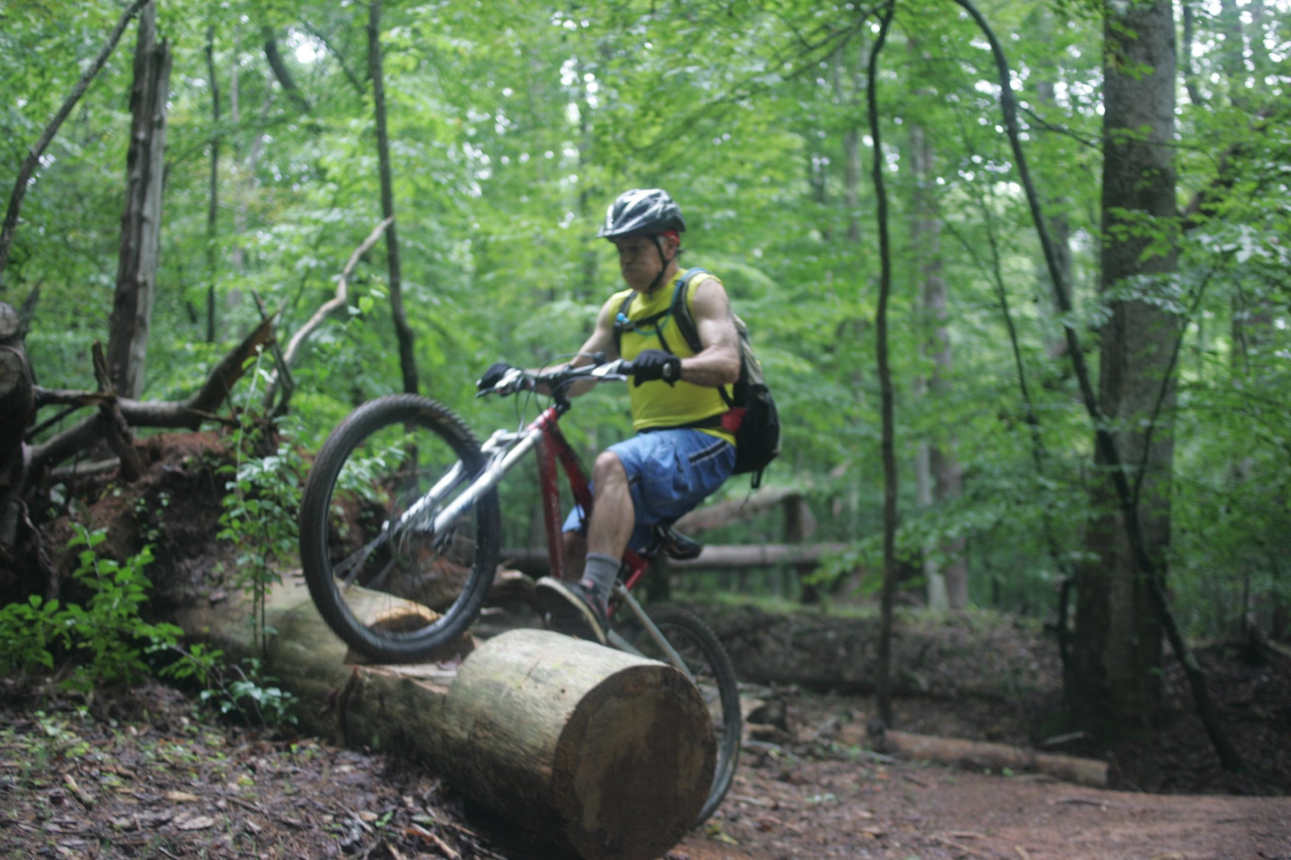 A cyclist in a yellow shirt and blue shorts performs a trick on a mountain bike, balancing on a fallen log in a lush green forest. Owls Roost (Bur-Mil Park) mountain bike trail.