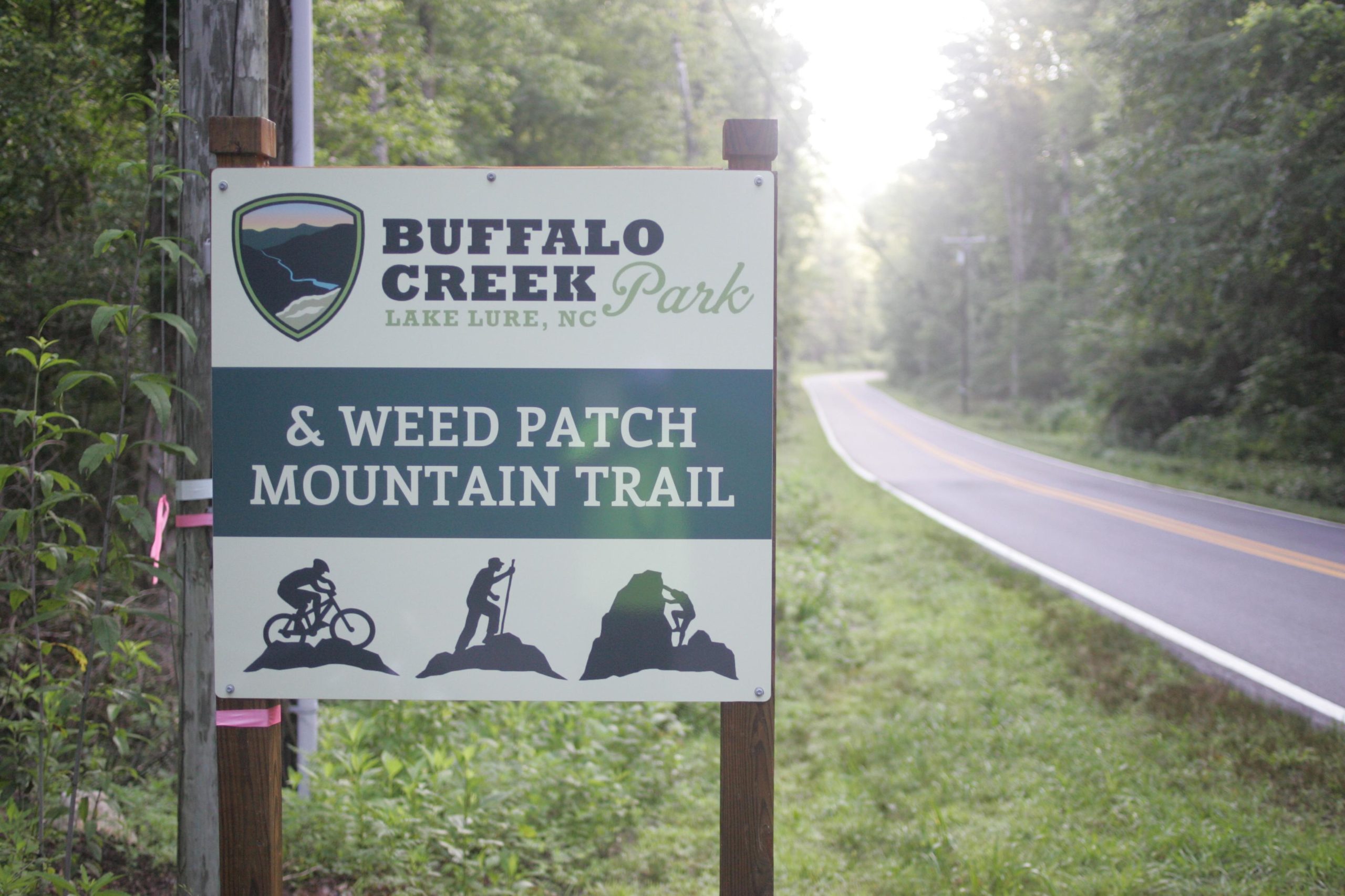 Sign for Buffalo Creek Park and Weed Patch Mountain Trail in Lake Lure, North Carolina, with a scenic road visible in the background surrounded by trees. Weed Patch Mountain Trail mountain bike trail.