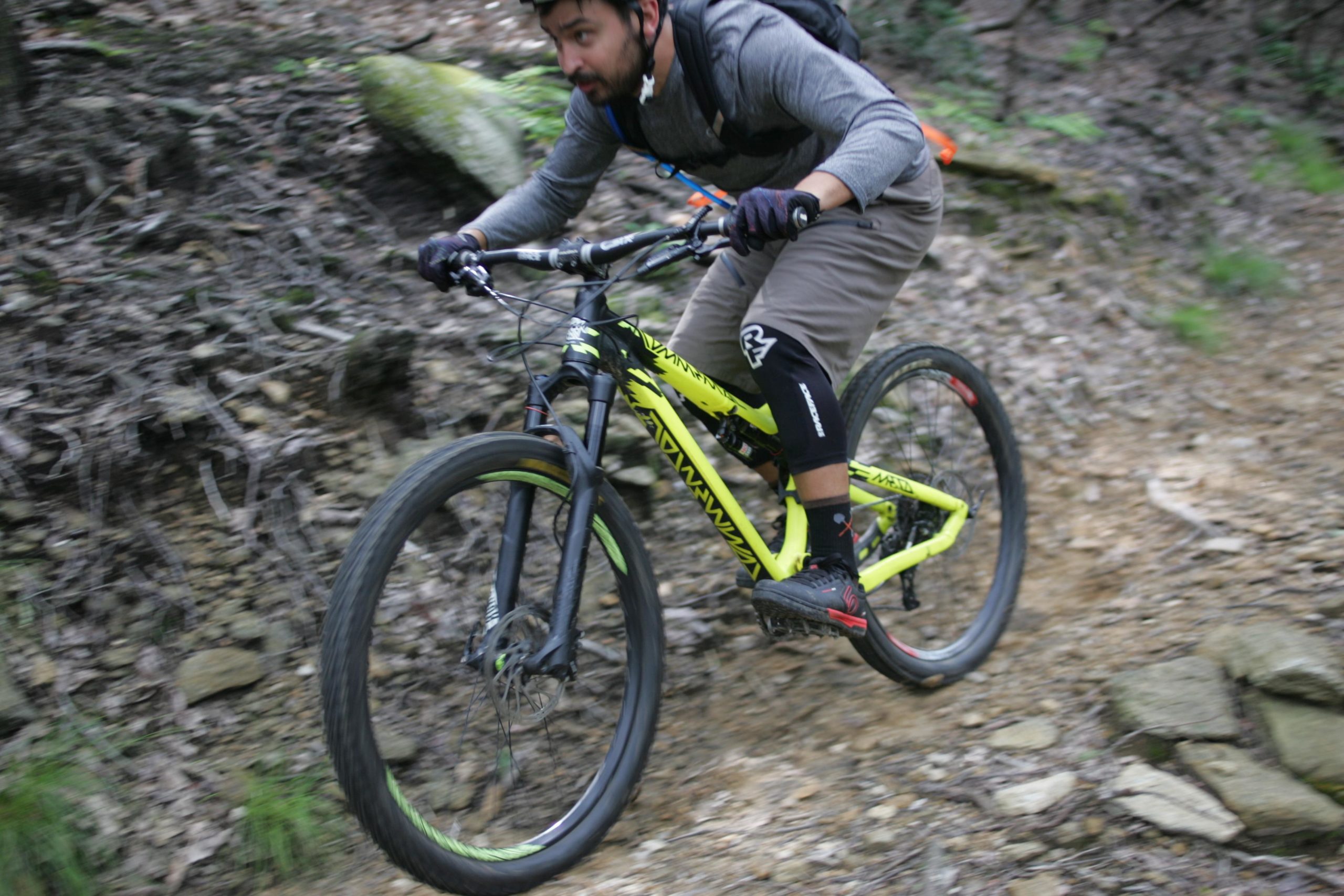 A mountain biker in motion navigating a rugged, wooded trail on a bright yellow bike. The rider is wearing a helmet and gloves, dressed in a gray long-sleeve shirt and tan shorts, showing focus and determination as they conquer the rocky terrain. Weed Patch Mountain Trail mountain bike trail.