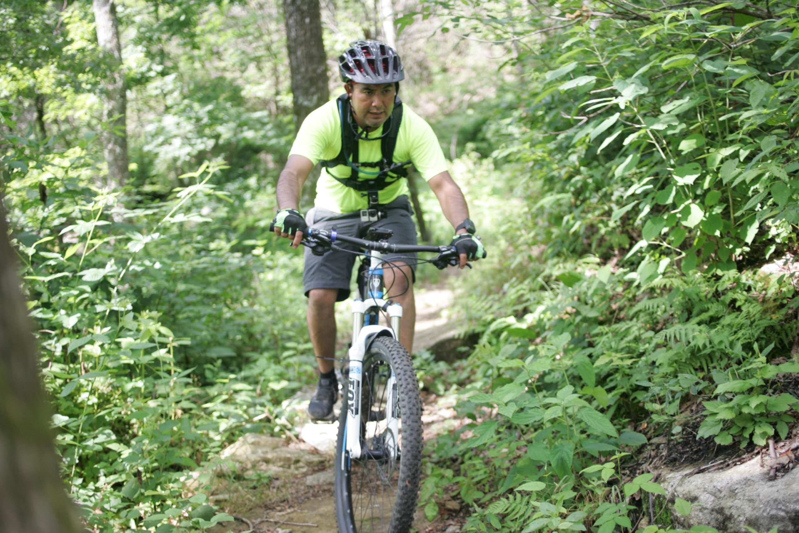 A cyclist wearing a bright yellow shirt and helmet rides a mountain bike on a narrow, rocky trail surrounded by lush greenery and trees. Weed Patch Mountain Trail mountain bike trail.