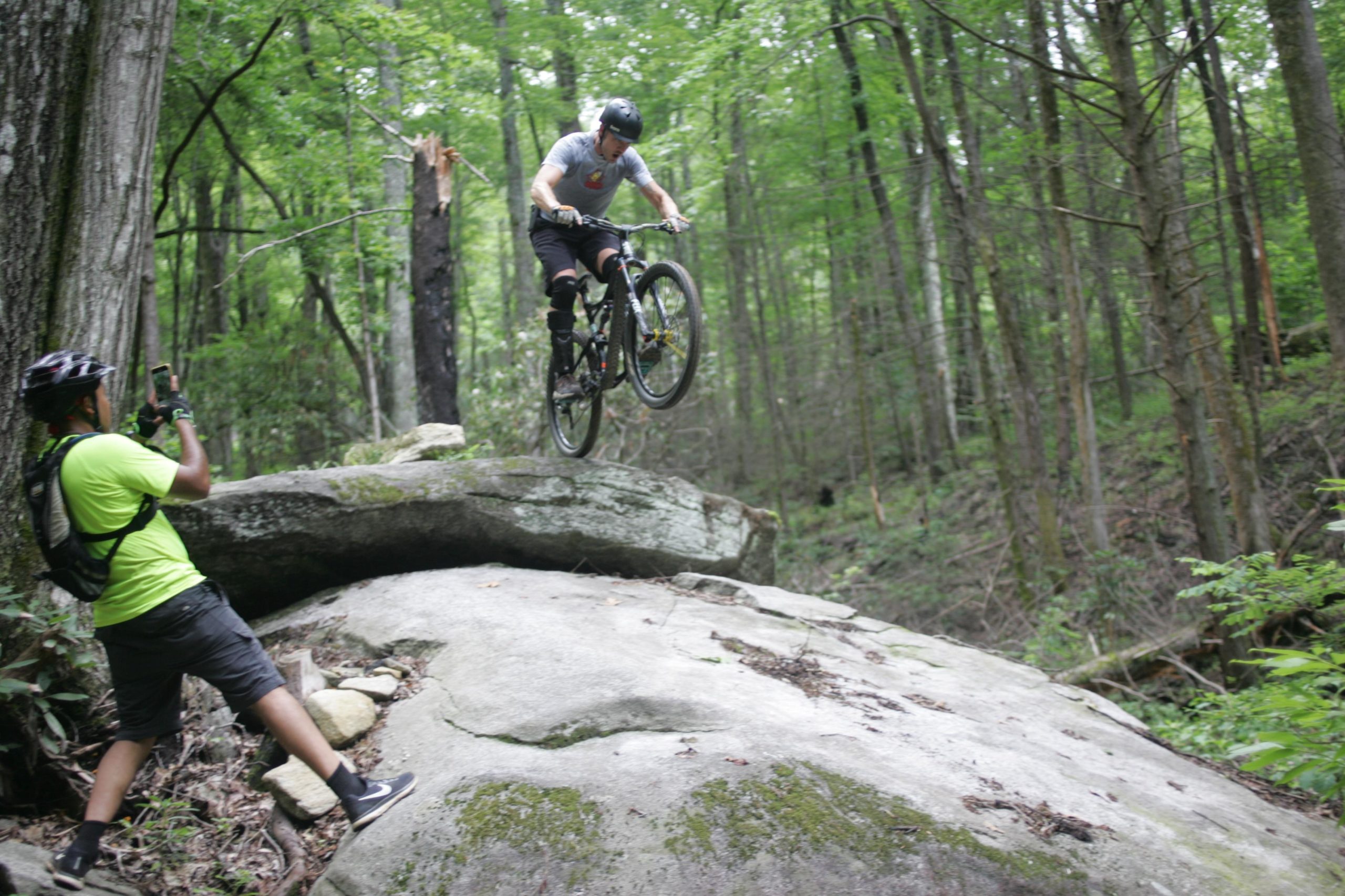 A mountain biker performing a jump over a large rock in a forested area, while another person is taking a photo of the action. The scene is surrounded by lush green trees and underbrush. Weed Patch Mountain Trail mountain bike trail.