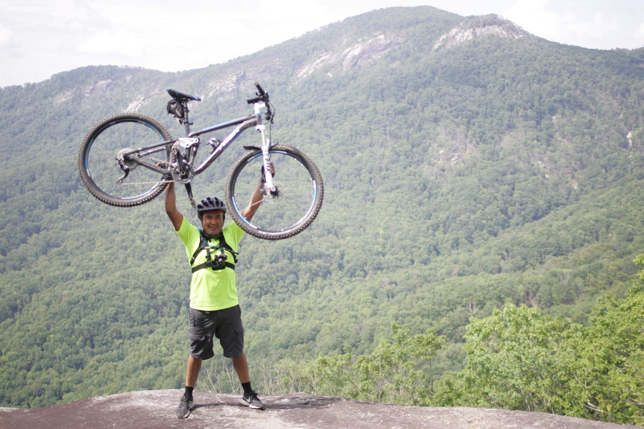 A person in a bright yellow shirt is triumphantly holding a mountain bike above their head while standing on a rocky outcrop, with a lush green forested mountain range in the background. The scene captures a sense of achievement and joy in an outdoor setting. Weed Patch Mountain Trail mountain bike trail.
