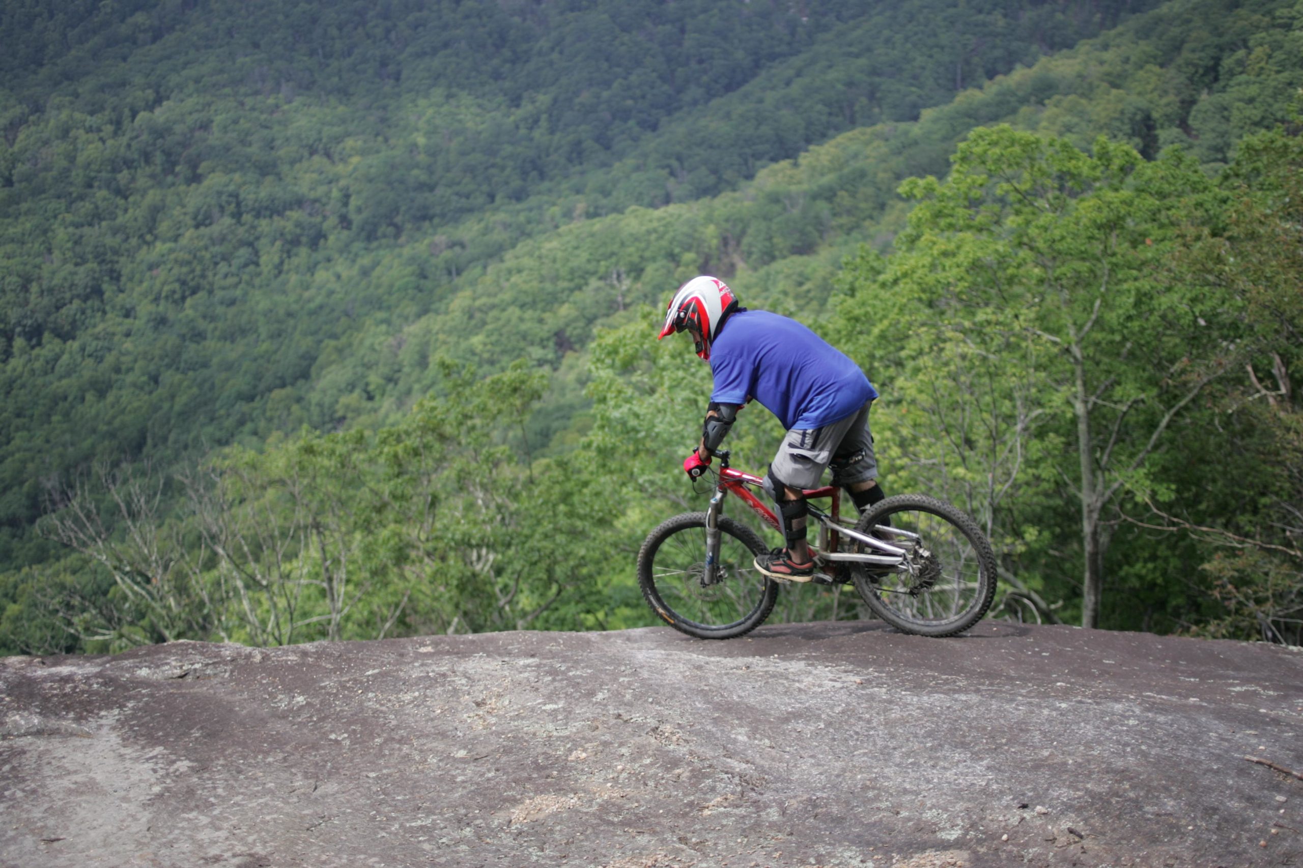 A person wearing a helmet and protective gear rides a mountain bike along the edge of a rocky surface, with a lush green forest and rolling hills in the background. Weed Patch Mountain Trail mountain bike trail.