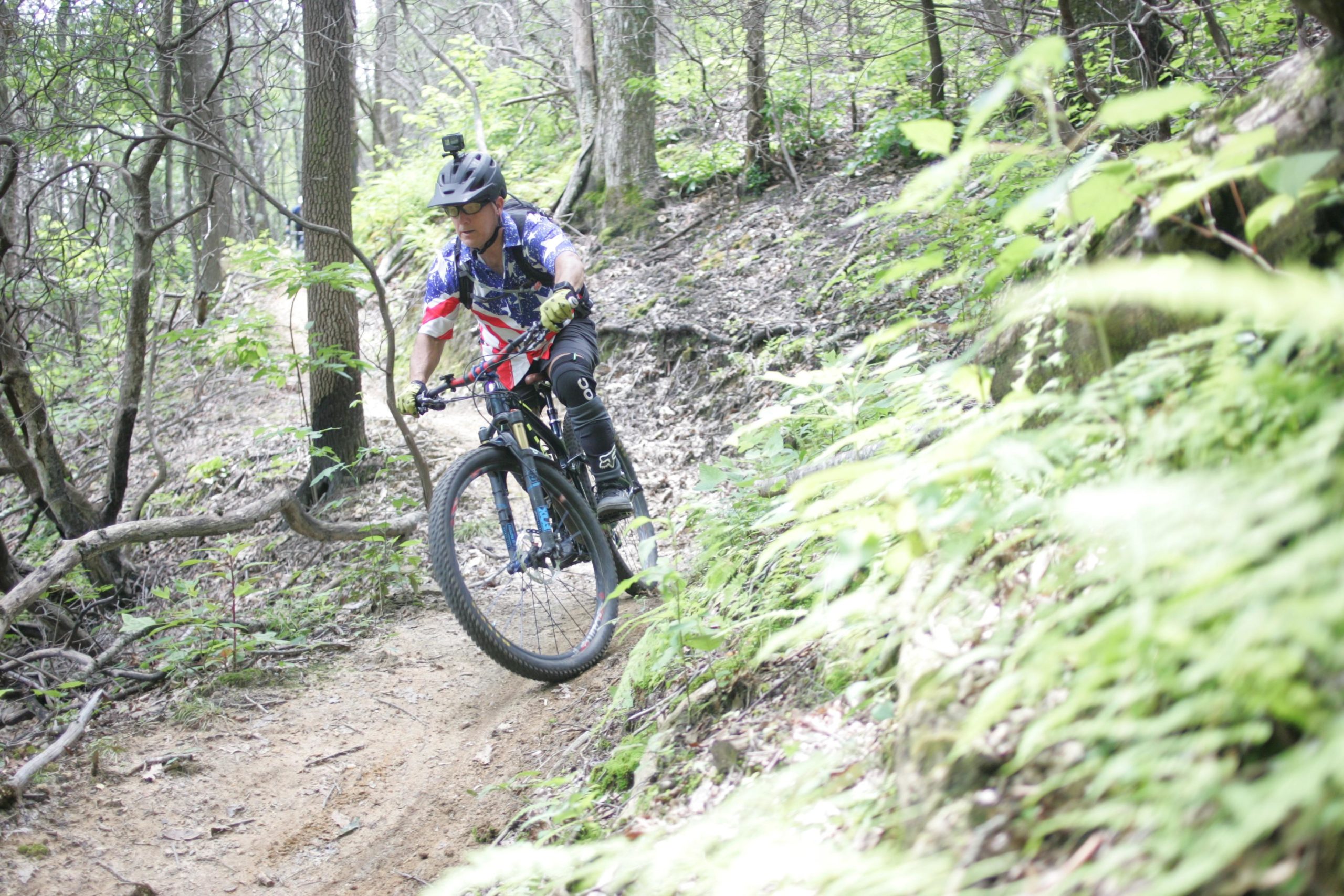 A cyclist wearing a helmet and colorful jersey navigates a trail through a wooded area, surrounded by greenery and tree trunks. The path is narrow and winding, with the rider leaning into a turn on a mountain bike. A camera is mounted on the cyclist's helmet, capturing the activity. Weed Patch Mountain Trail mountain bike trail.