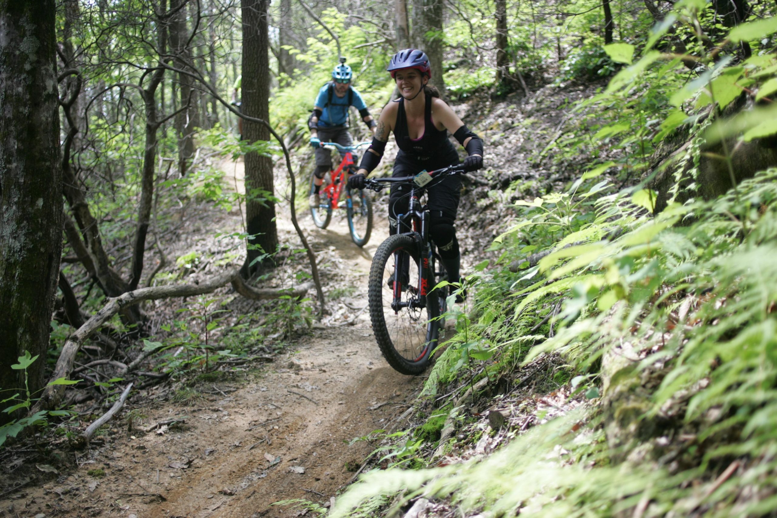 A woman rides a mountain bike on a dirt trail surrounded by lush greenery and trees. Another cyclist is visible in the background, navigating the trail. The scene captures the excitement of mountain biking in a forested area. Weed Patch Mountain Trail mountain bike trail.