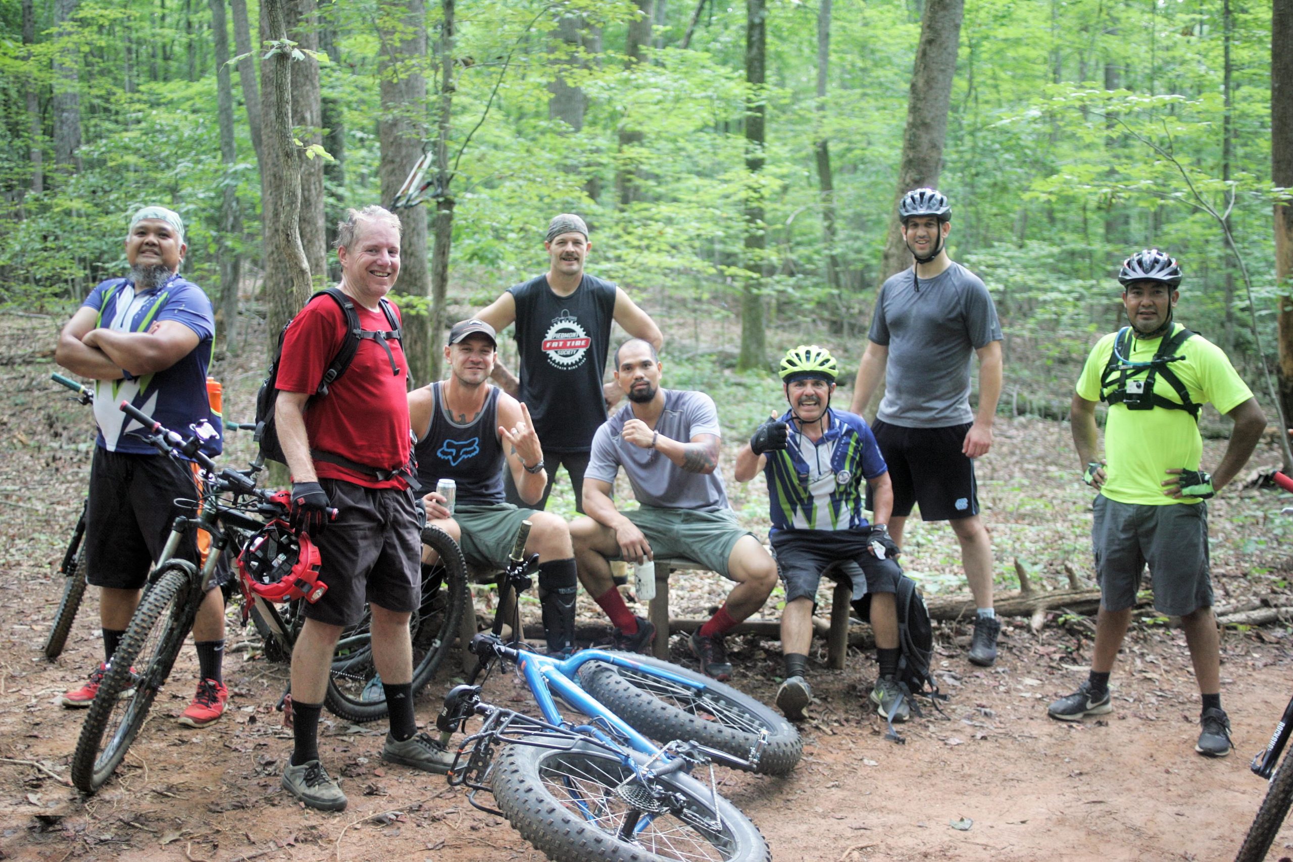 A group of eight men posing for a photo in a forested area, surrounded by trees. They are dressed in casual outdoor gear and mountain biking attire, with various bicycles leaned against nearby trees. The men are smiling and appear to be enjoying their time together after biking. The scene conveys a sense of camaraderie and outdoor adventure. Salem Lake mountain bike trail.