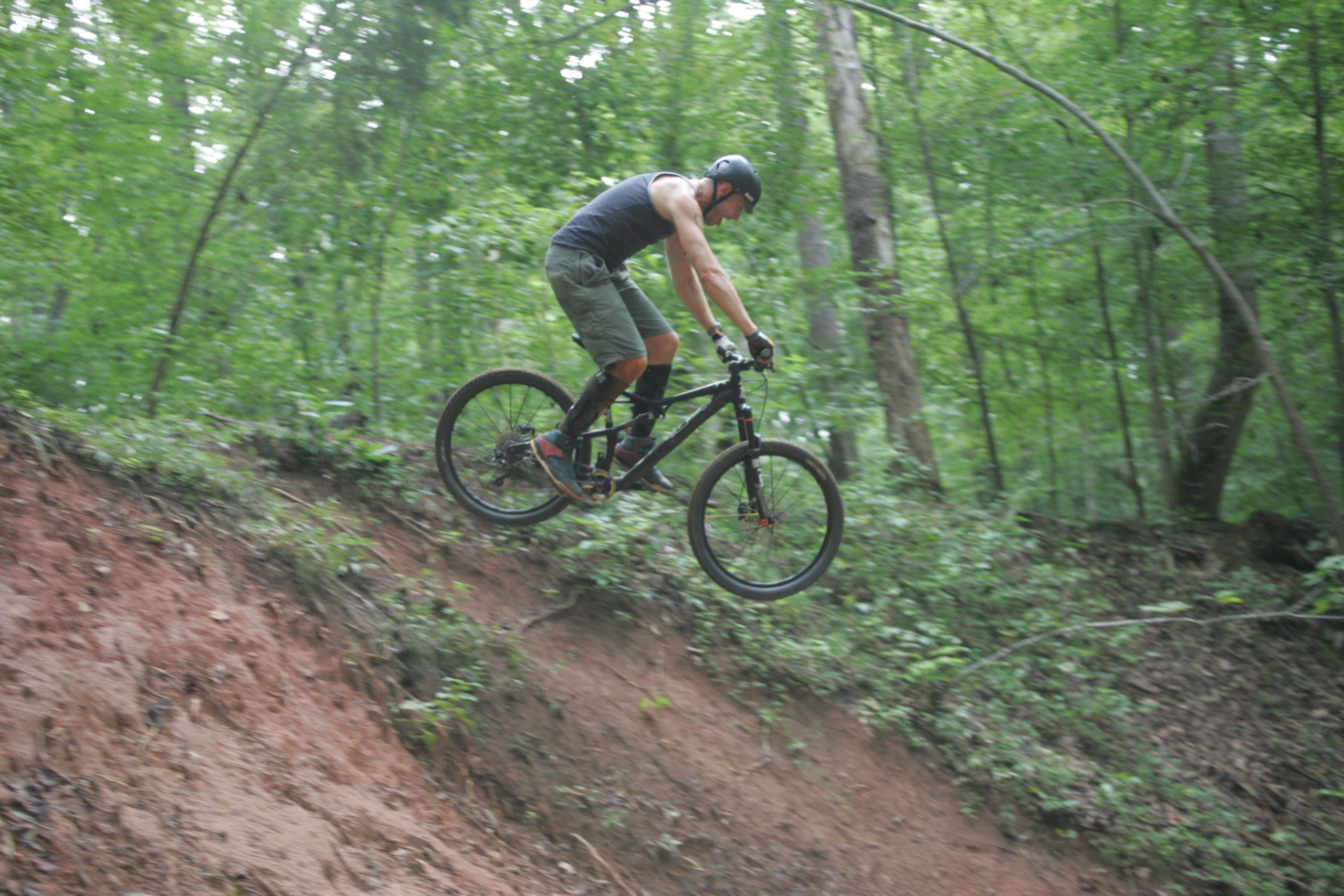 A mountain biker performing a jump over a dirt ramp in a lush green forest. The rider is wearing a helmet and protective gear, capturing a moment of action and excitement. Salem Lake mountain bike trail.