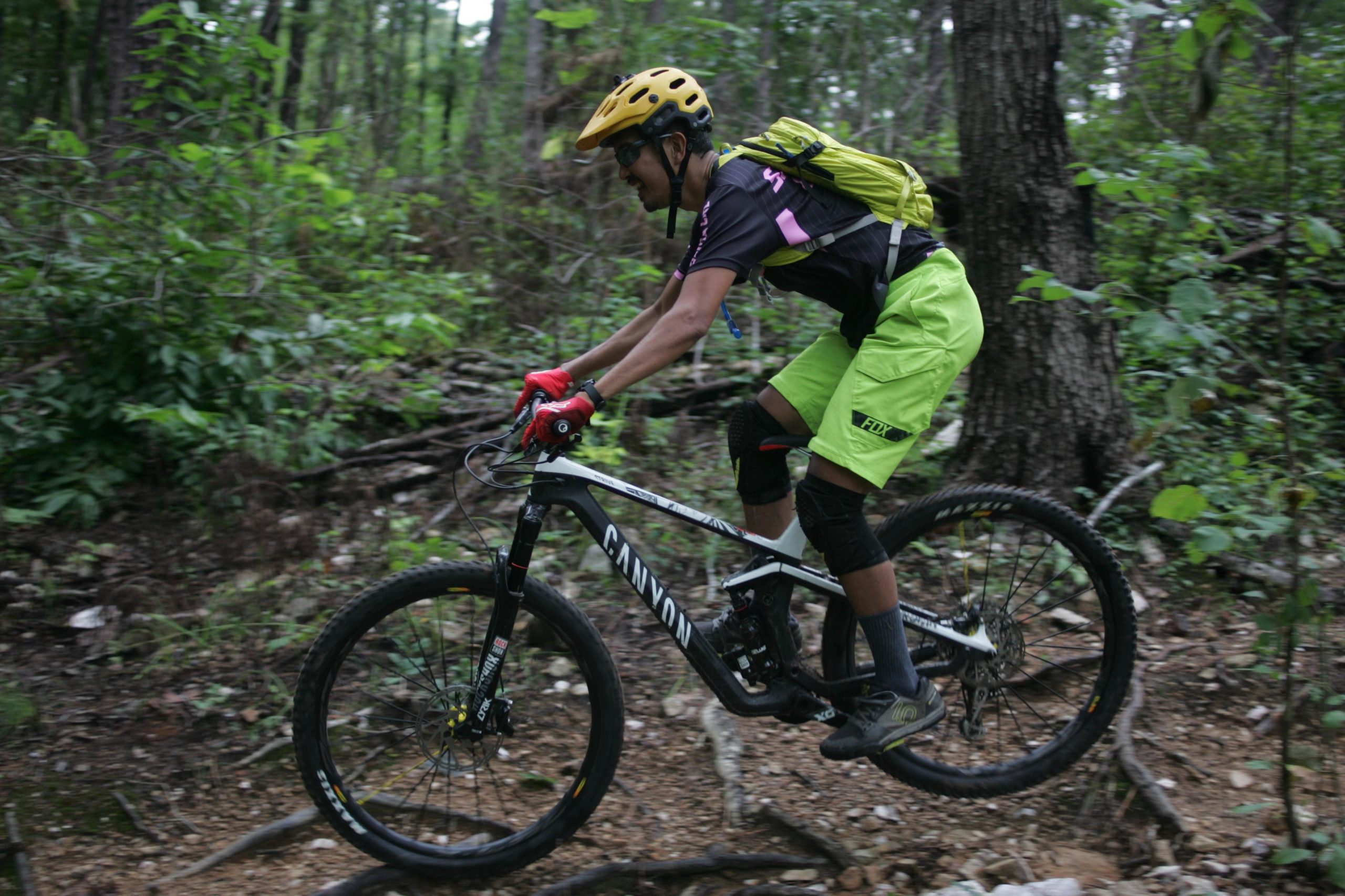 A person riding a mountain bike on a rugged forest trail, wearing a helmet and bright yellow shorts, with a green backpack. The background features trees and foliage, indicating a natural outdoor setting. New Light Trails mountain bike trail.