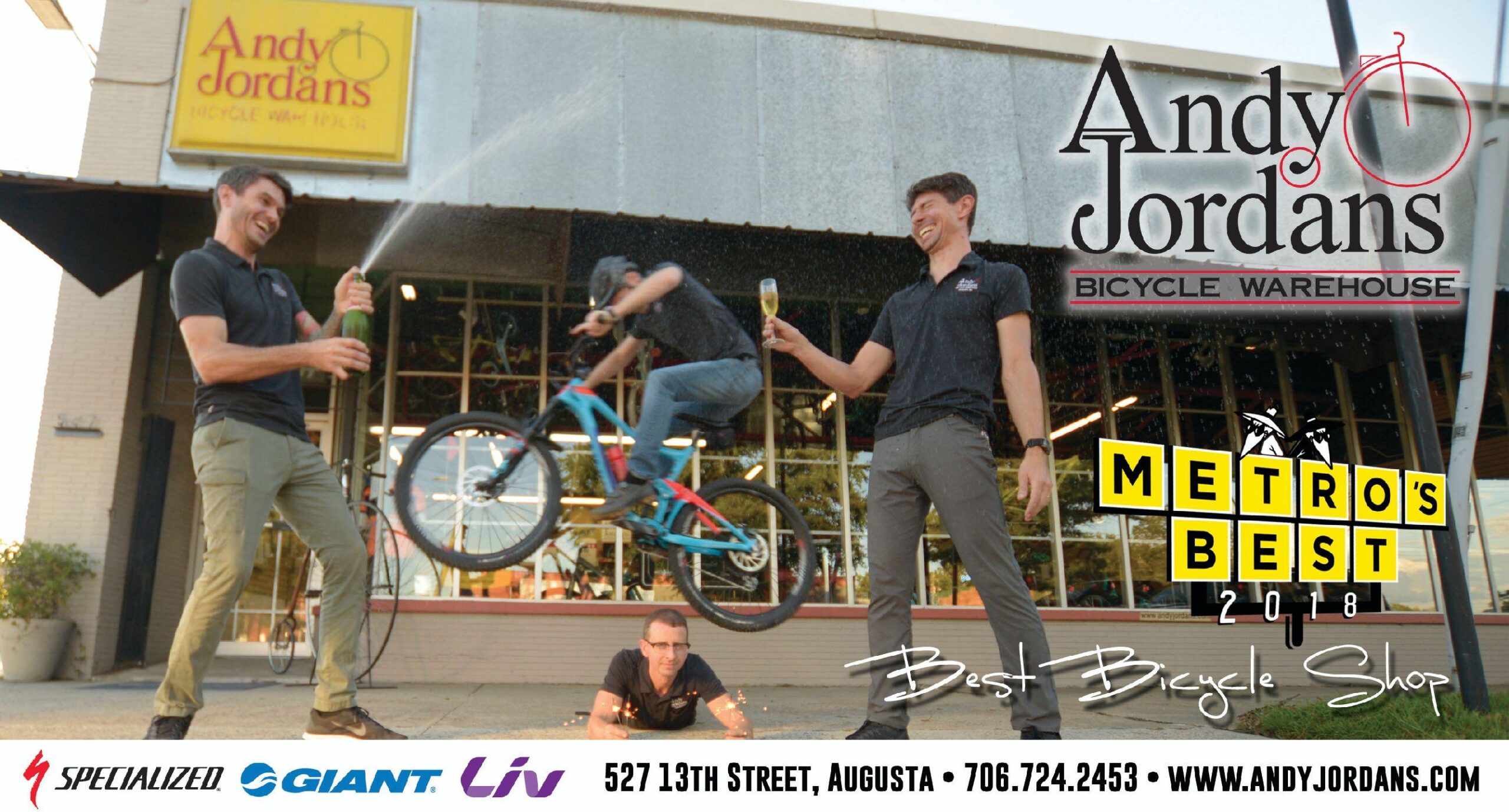 A celebration outside Andy Jordan's Bicycle Warehouse, featuring three men: one is popping a champagne bottle, another is performing a trick on a bicycle mid-air, and the third is lying on the ground with sparkles around him. The store sign and a "Metro's Best 2018" award for Best Bicycle Shop are visible in the background.