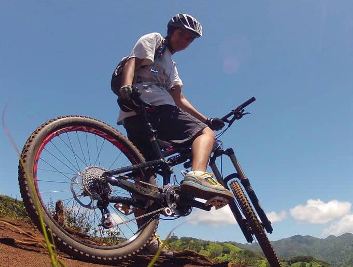 A mountain biker wearing a helmet and casual sports attire is balancing on a bike on a dirt trail. The background features a clear blue sky and green hills, indicating an outdoor cycling environment.