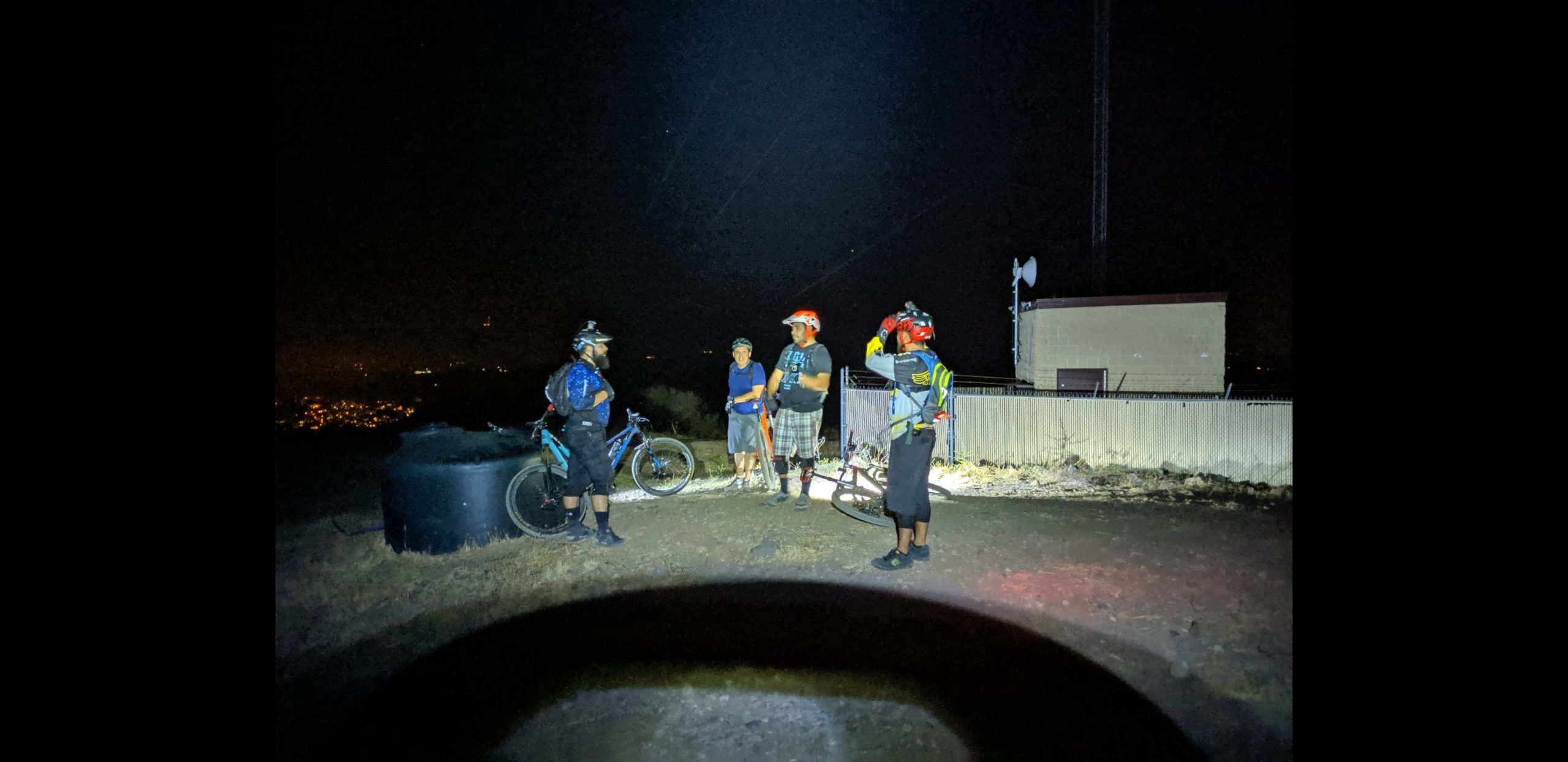A group of four mountain bikers gathered at night on a hillside, illuminated by a bright light. They are wearing helmets and biking gear, standing next to their bicycles. In the background, a building and a fence are visible, along with city lights twinkling in the distance. Hill Canyon mountain bike trail.