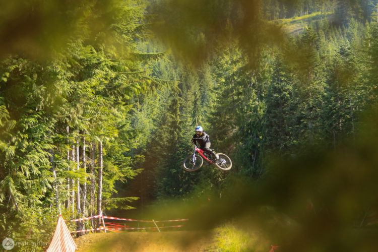 A mountain biker in mid-air performing a jump over a dirt track, surrounded by lush green trees in a forest setting. Red and white caution tape indicates the biking area below.