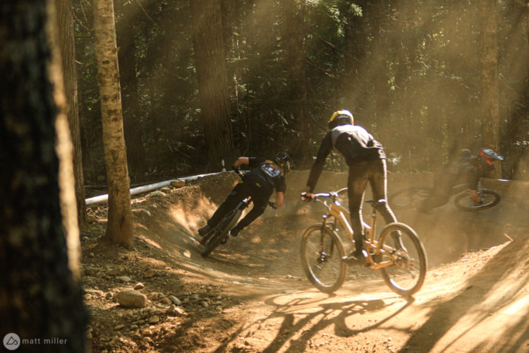 Two mountain bikers racing along a dirt trail in a forest, surrounded by trees and dust particles illuminated by sunlight. One biker is leaning into a turn while the other is slightly behind, both wearing helmets and riding in a dynamic pose.