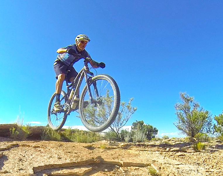 A young cyclist performing a jump on a mountain bike over rocky terrain under a blue sky, with shrubs in the background. White Ridge Bike Trails mountain bike trail.