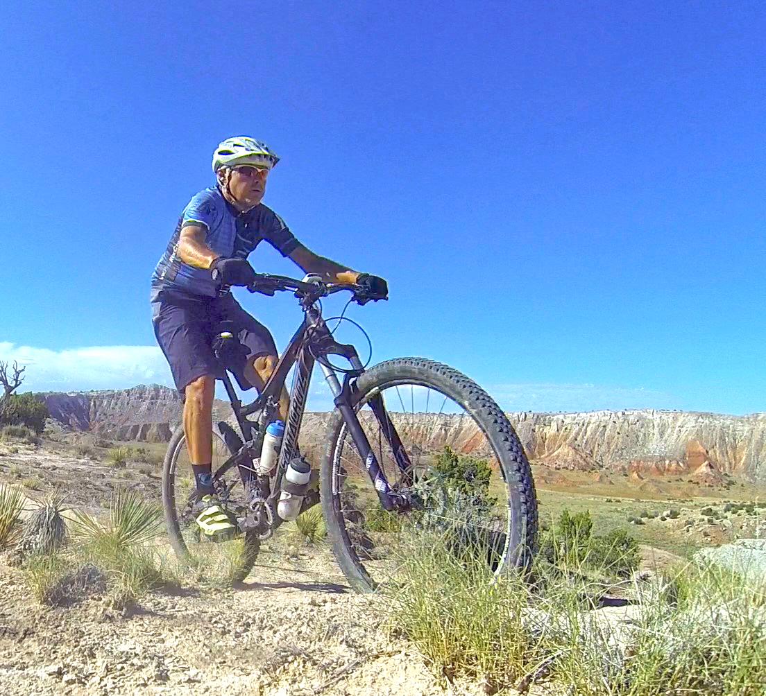 A person riding a mountain bike on a dirt trail, surrounded by rocky terrain and low vegetation, under a clear blue sky. The landscape features colorful rock formations in the background, suggesting a scenic outdoor environment. White Ridge Bike Trails mountain bike trail.