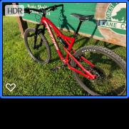 Fezzari Abajo Peak: A red mountain bike is positioned on green grass in front of a sign with the text "Kane Co." The bike features a sturdy frame, wide tires, and a suspension system, indicating it is designed for off-road trails.