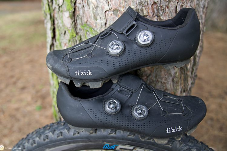A pair of black cycling shoes from the brand fi'zi:k, featuring a sleek design with two circular dial closures for adjustments. The shoes are positioned against a tree trunk and have some dirt on them, indicating outdoor use. The sole is equipped with grips suitable for mountain biking.
