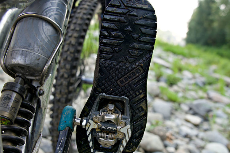 Close-up view of a mountain bike pedal attached to a rider's shoe, featuring a textured sole. The background shows a rocky surface with greenery, indicating an outdoor terrain suitable for biking.