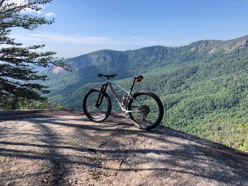 A mountain bike positioned on a rocky ledge overlooking a lush green valley and distant mountains under a clear blue sky. Weed Patch Mountain Trail mountain bike trail.